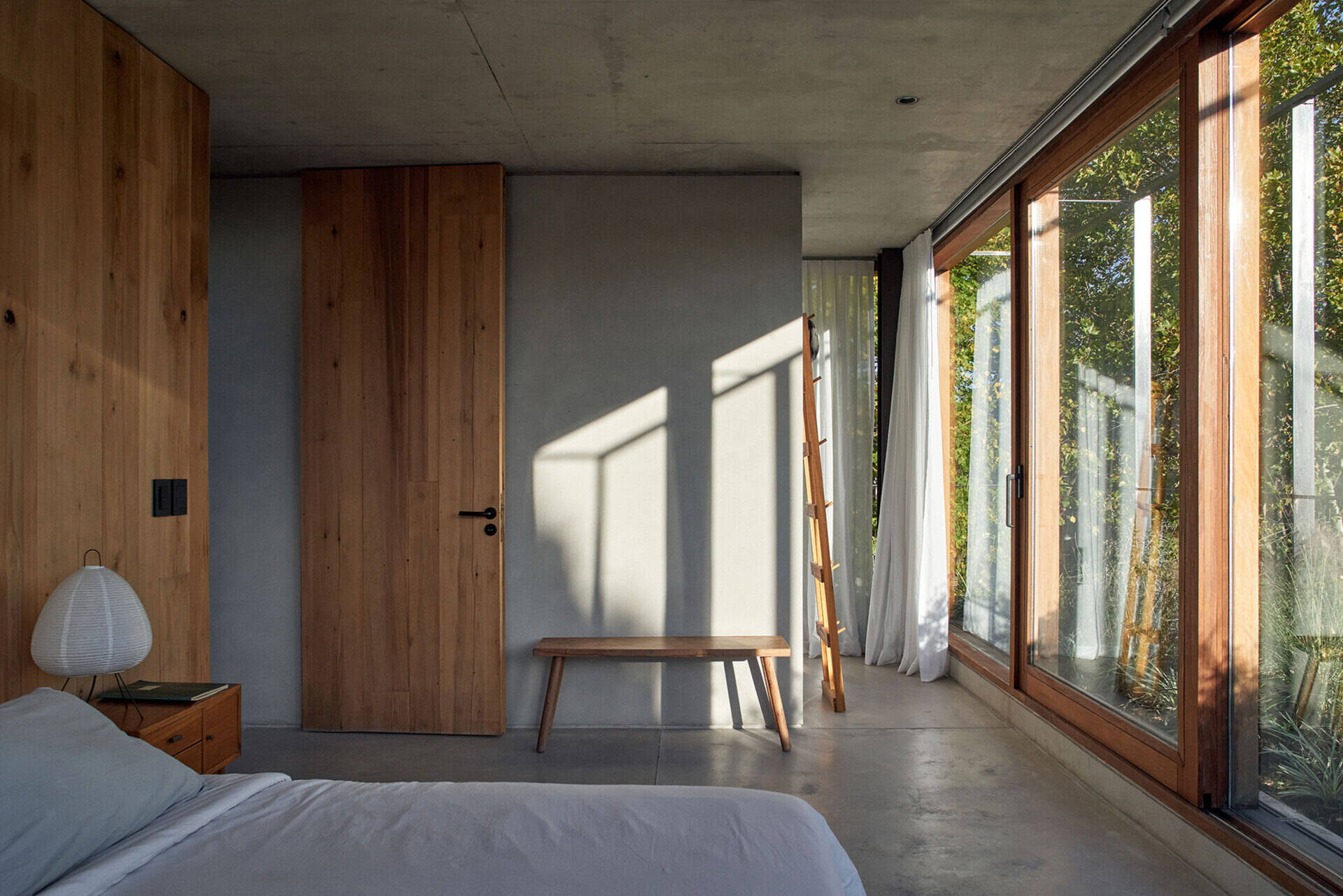 A bedroom lined with floor-to-ceiling windows brings in natural light while framing views of the surrounding trees.