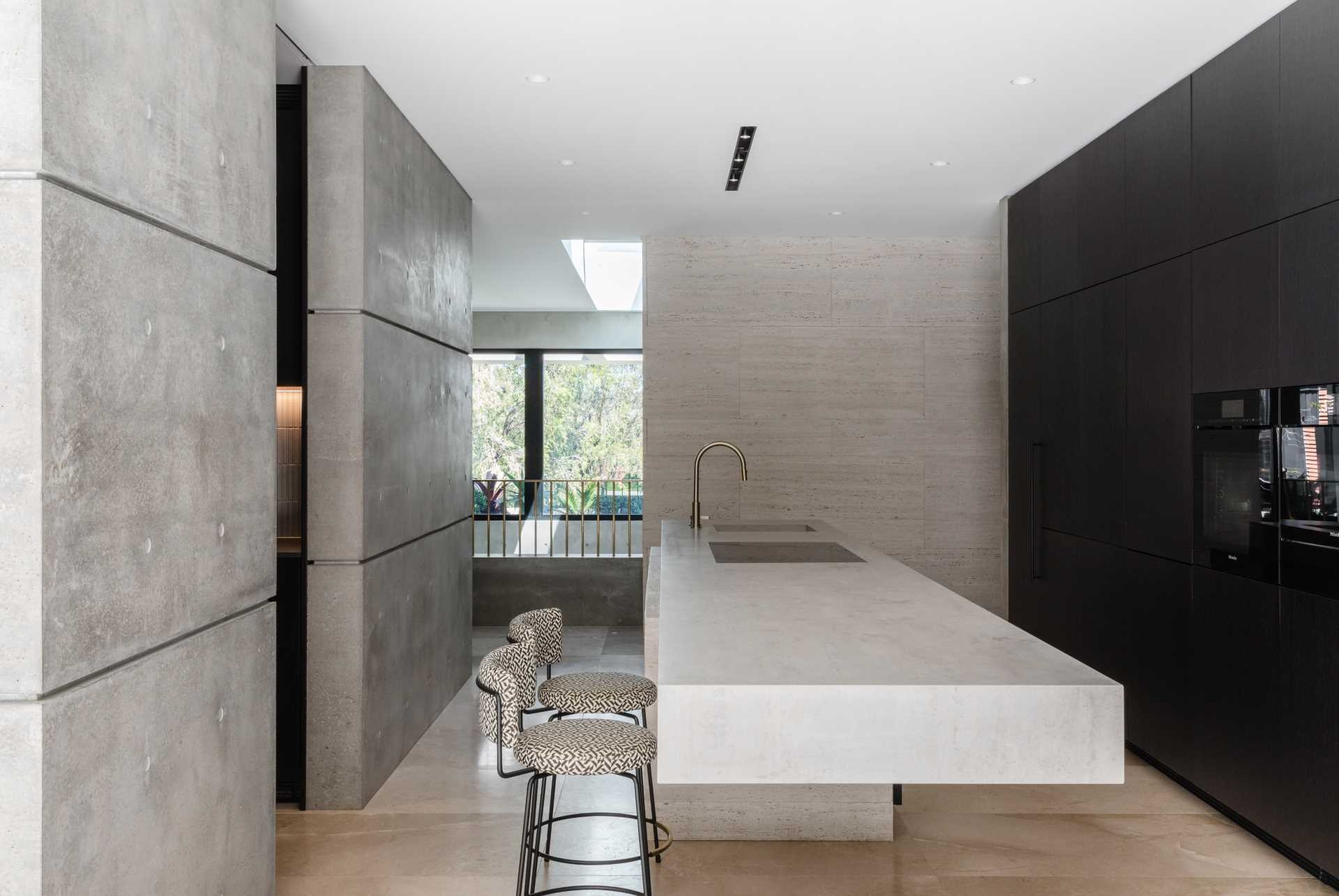 The minimalist kitchen in this Sydney home features a dramatic cantilevered island, black cabinetry, concrete walls, limestone floors, and travertine finishes that create a bold yet refined modern aesthetic.