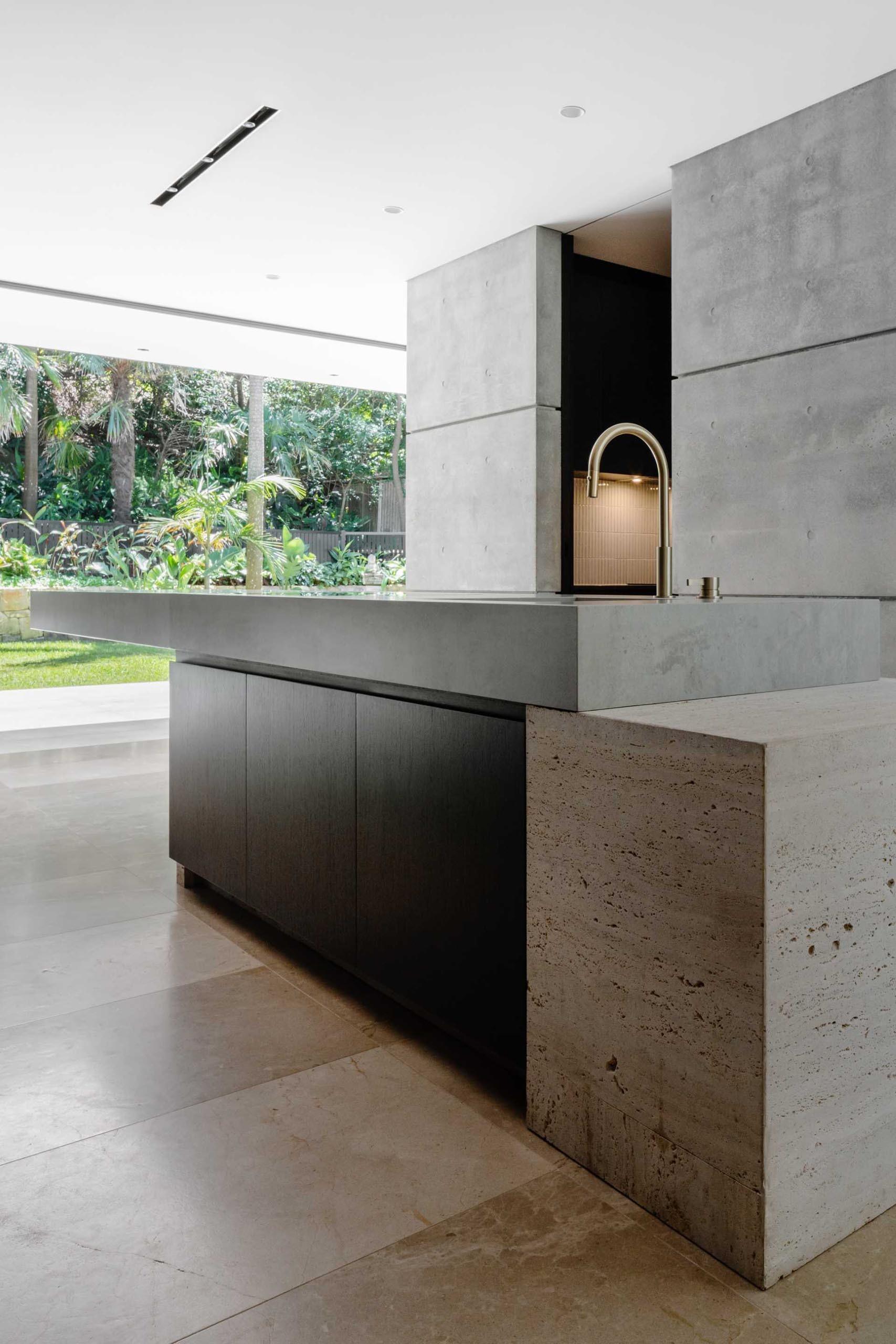 The minimalist kitchen in this Sydney home features a dramatic cantilevered island, black cabinetry, concrete walls, limestone floors, and travertine finishes that create a bold yet refined modern aesthetic.