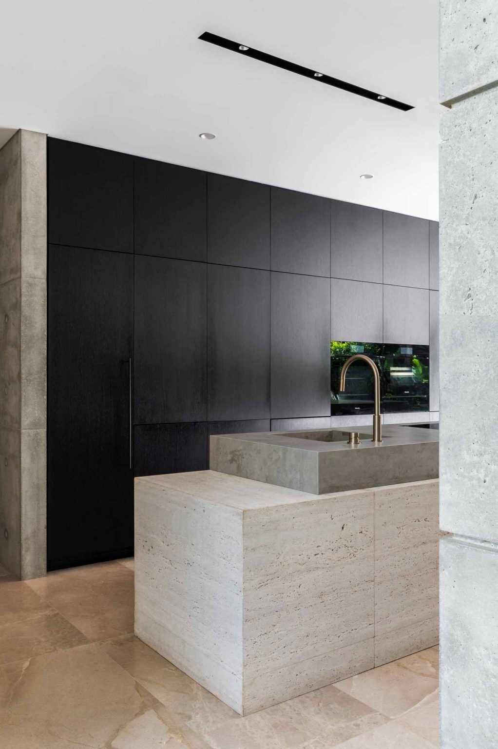 The minimalist kitchen in this Sydney home features a dramatic cantilevered island, black cabinetry, concrete walls, limestone floors, and travertine finishes that create a bold yet refined modern aesthetic.