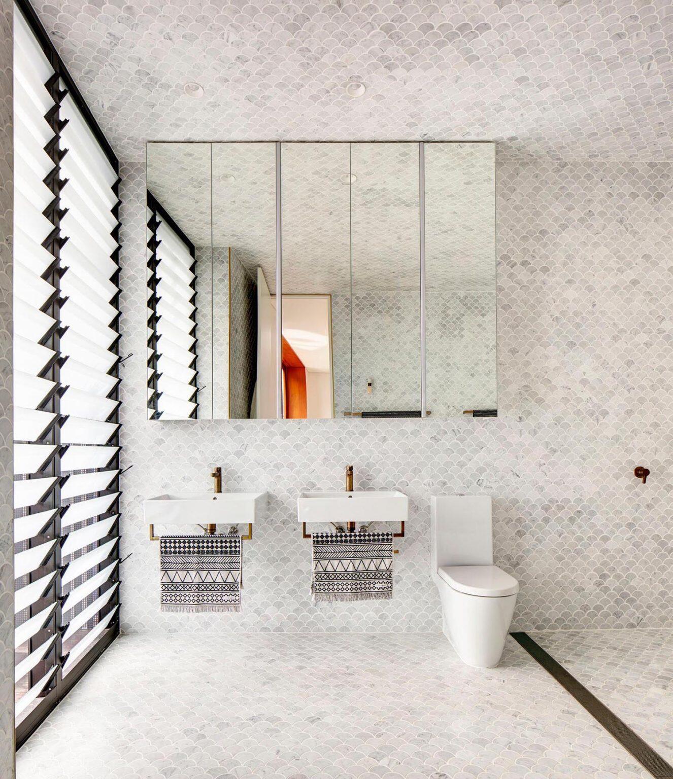 The main ensuite is wrapped in light grey fan-shaped tiles that extend across ceilings and tall walls, paired with brass fixtures that introduce warmth against the cool surface.
