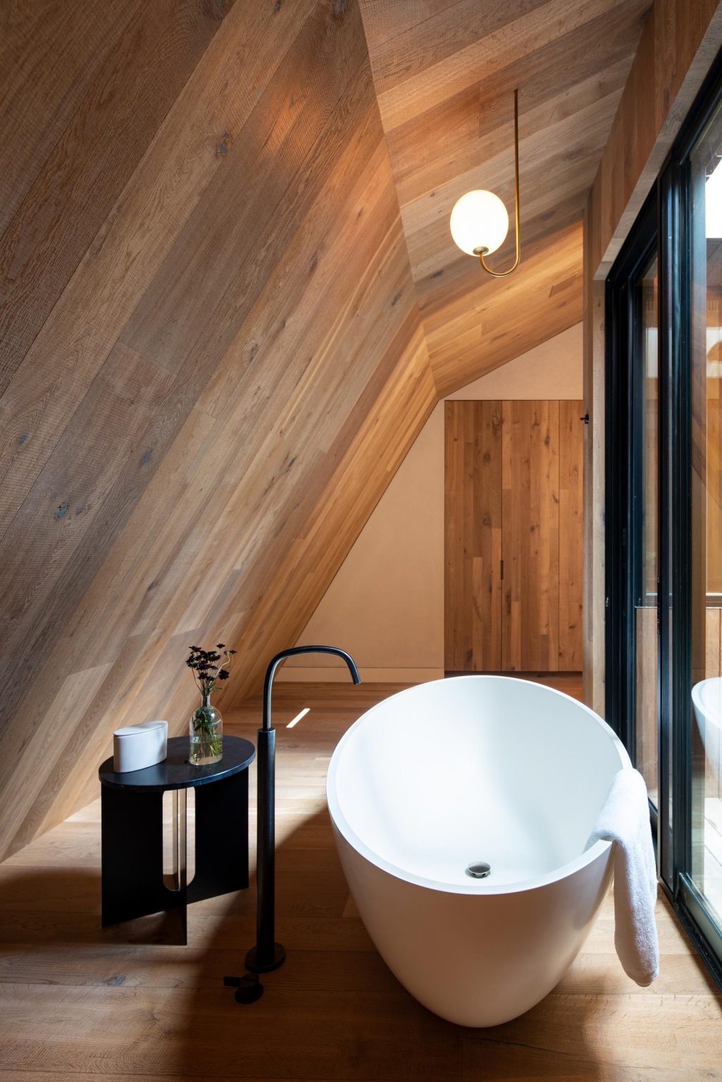 The en-suite bathroom continues the warm material palette with a wood vanity that blends seamlessly into the wall behind it. Vertical detailing emphasizes the height of the ceiling, while a freestanding bathtub and minimalist black faucet complete the serene space.