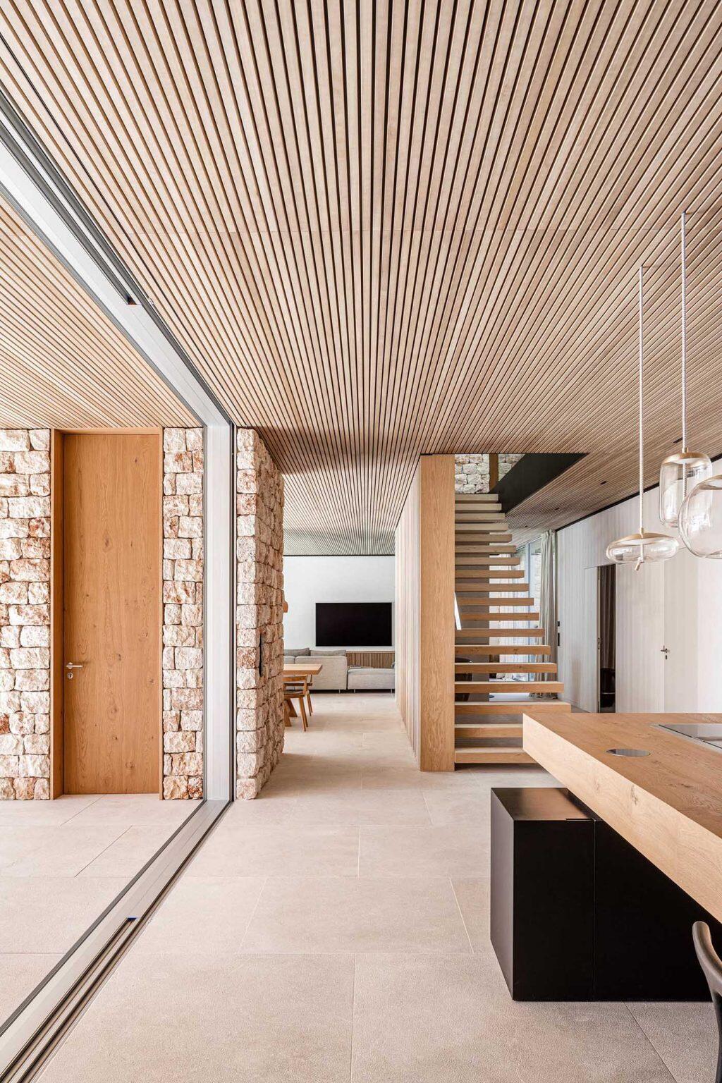 A staircase leads down to the main living spaces below. Vertical wooden slats positioned beside the stairs create a striking architectural feature that acts almost like the structural skeleton of the interior. Natural light filters through the space, highlighting the textures of stone, wood, lime plaster, and concrete used throughout the home.