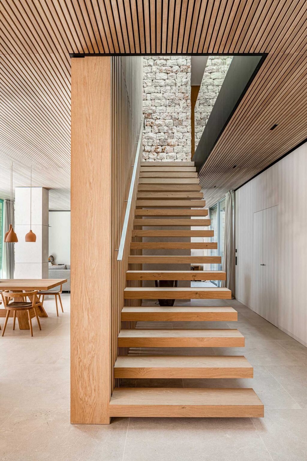 A staircase leads down to the main living spaces below. Vertical wooden slats positioned beside the stairs create a striking architectural feature that acts almost like the structural skeleton of the interior. Natural light filters through the space, highlighting the textures of stone, wood, lime plaster, and concrete used throughout the home.