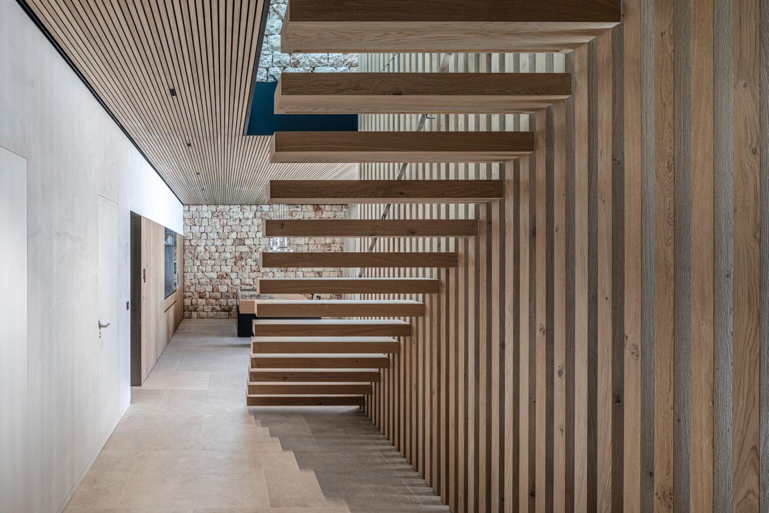A staircase leads down to the main living spaces below. Vertical wooden slats positioned beside the stairs create a striking architectural feature that acts almost like the structural skeleton of the interior. Natural light filters through the space, highlighting the textures of stone, wood, lime plaster, and concrete used throughout the home.