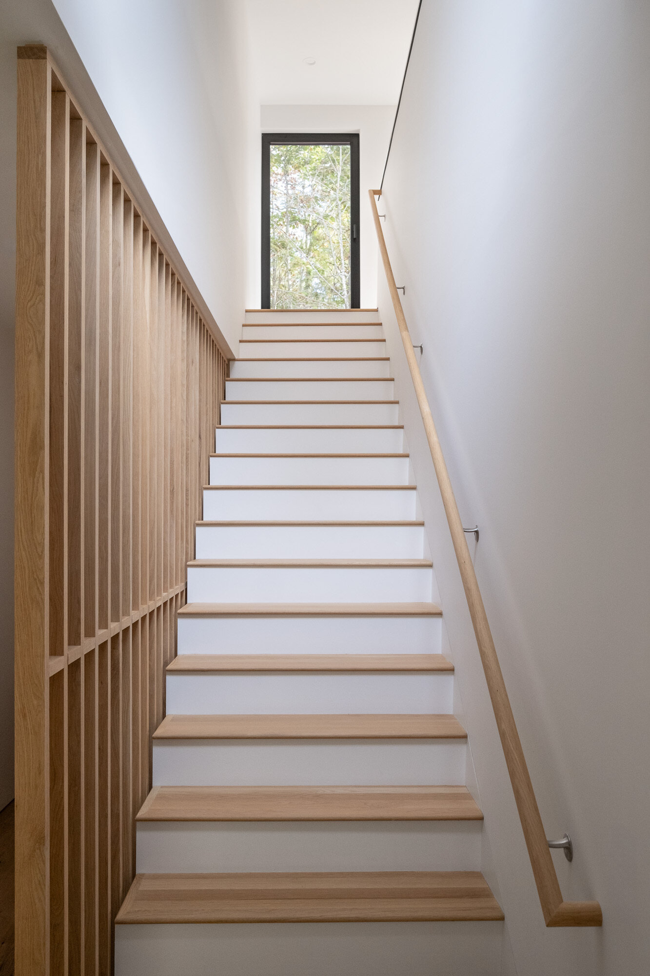 The wood and white staircase acts connects between levels, while natural light filtering through nearby glazing and the wood slat divider helps prevent the stair zone from feeling enclosed.