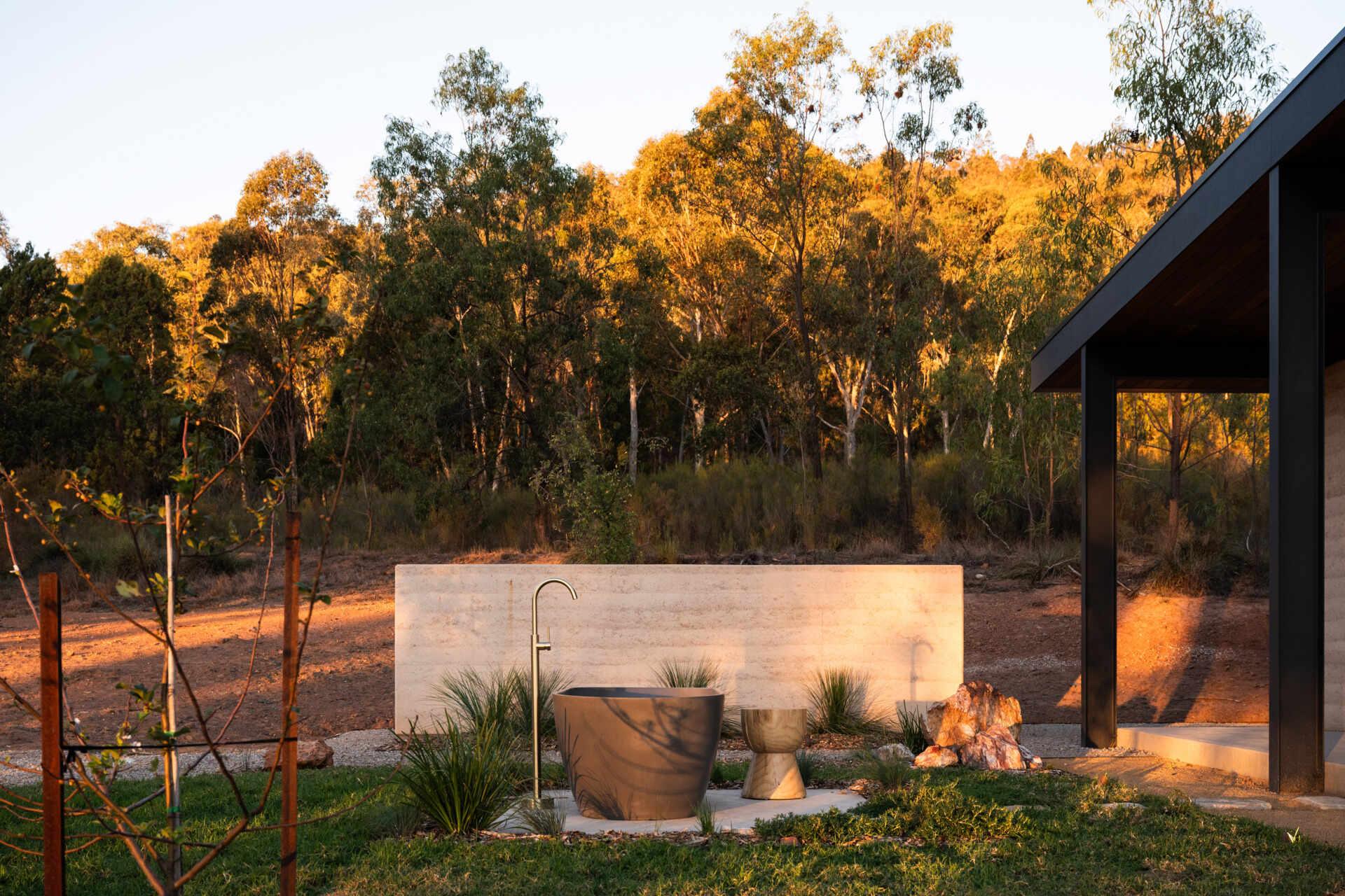 A contemporary off-grid cabin in Mudgee with an open-plan layout that combines the living area, bedroom, and kitchen into one light-filled space. Rammed earth walls, warm timber finishes, a minimalist bathroom, and an outdoor bathtub overlooking the landscape create a relaxing vineyard farm stay surrounded by Australian art and valley views.