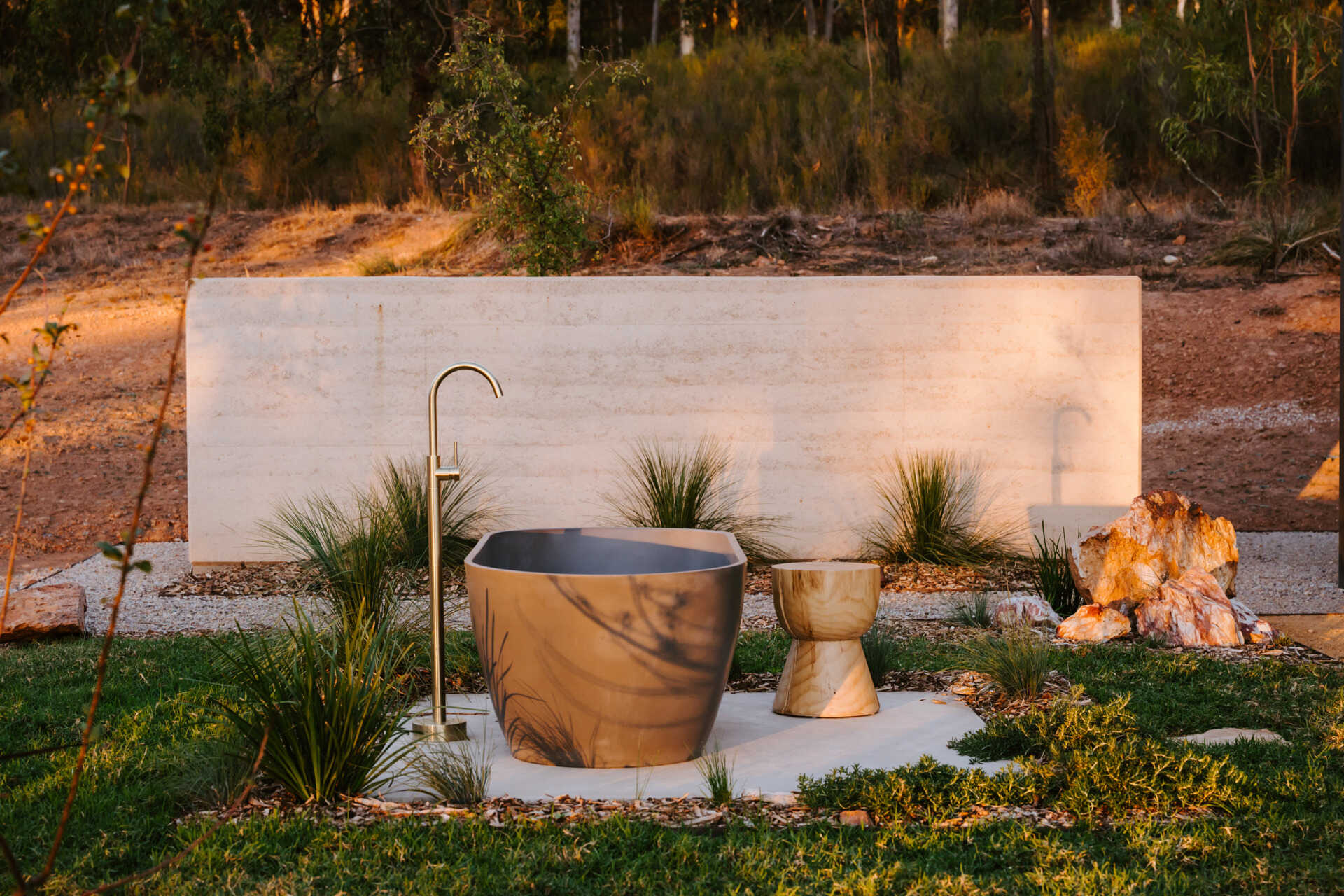 A contemporary off-grid cabin in Mudgee with an open-plan layout that combines the living area, bedroom, and kitchen into one light-filled space. Rammed earth walls, warm timber finishes, a minimalist bathroom, and an outdoor bathtub overlooking the landscape create a relaxing vineyard farm stay surrounded by Australian art and valley views.