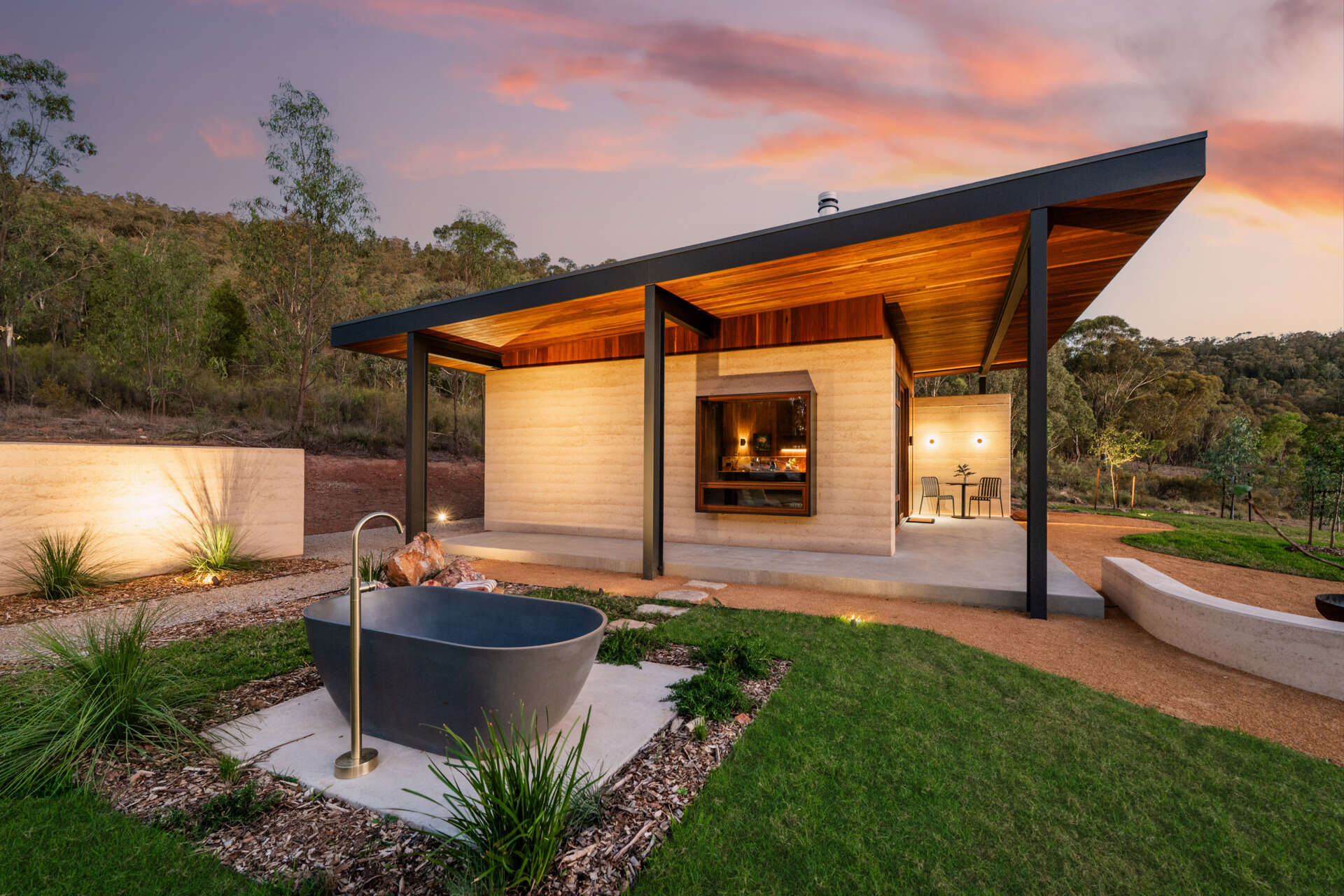A contemporary off-grid cabin in Mudgee with an open-plan layout that combines the living area, bedroom, and kitchen into one light-filled space. Rammed earth walls, warm timber finishes, a minimalist bathroom, and an outdoor bathtub overlooking the landscape create a relaxing vineyard farm stay surrounded by Australian art and valley views.