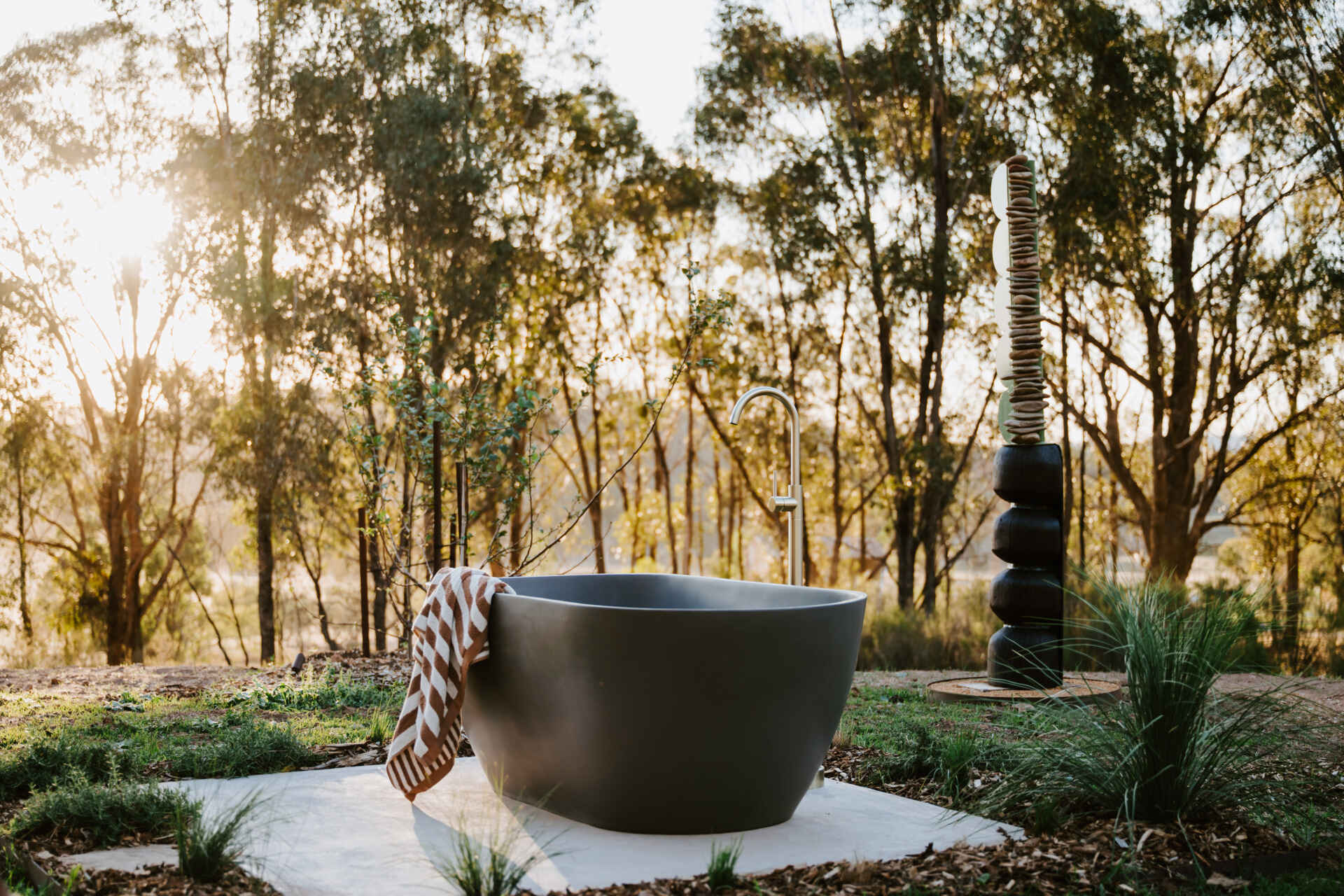 A contemporary off-grid cabin in Mudgee with an open-plan layout that combines the living area, bedroom, and kitchen into one light-filled space. Rammed earth walls, warm timber finishes, a minimalist bathroom, and an outdoor bathtub overlooking the landscape create a relaxing vineyard farm stay surrounded by Australian art and valley views.
