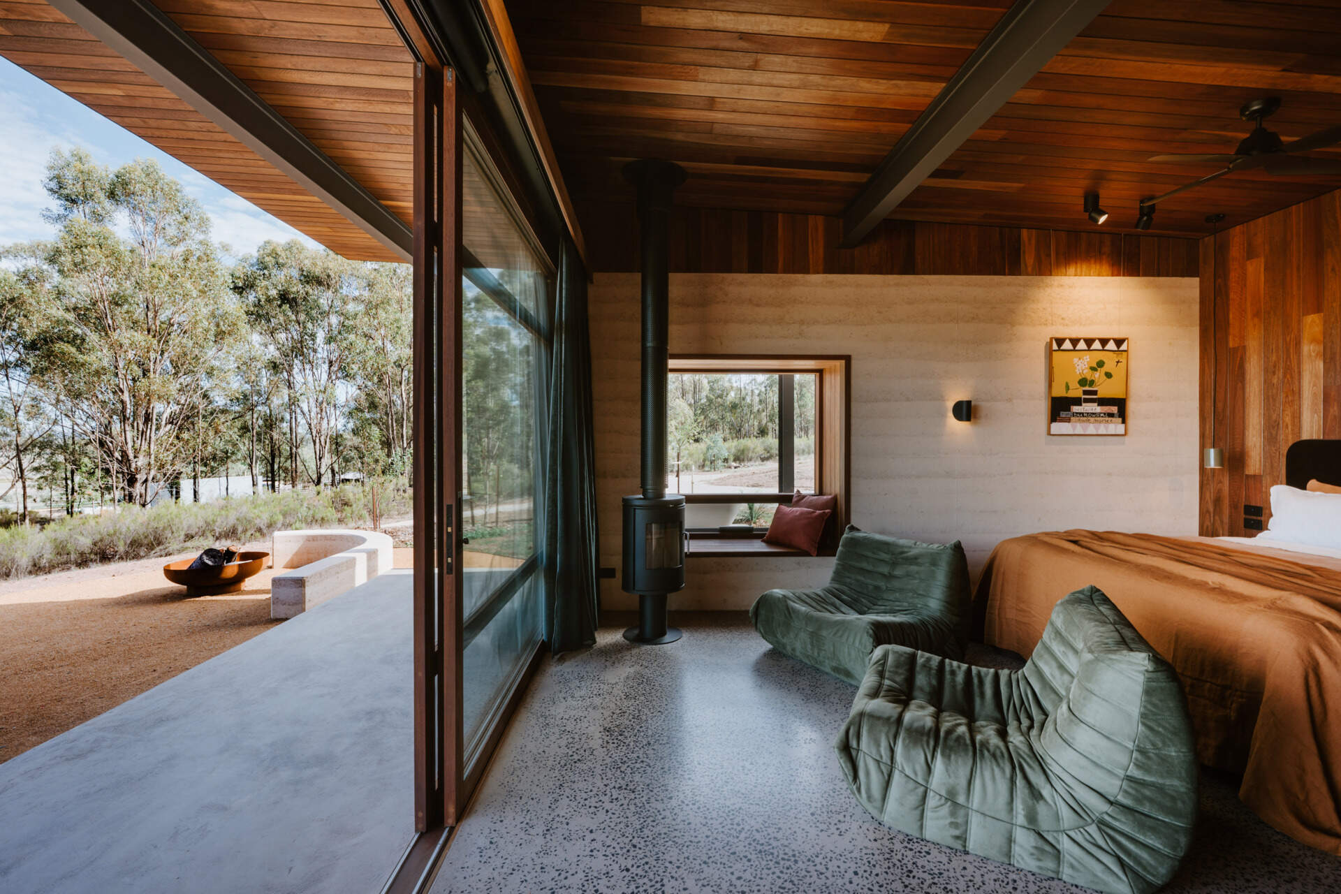 A contemporary off-grid cabin in Mudgee with an open-plan layout that combines the living area, bedroom, and kitchen into one light-filled space. Rammed earth walls, warm timber finishes, a minimalist bathroom, and an outdoor bathtub overlooking the landscape create a relaxing vineyard farm stay surrounded by Australian art and valley views.