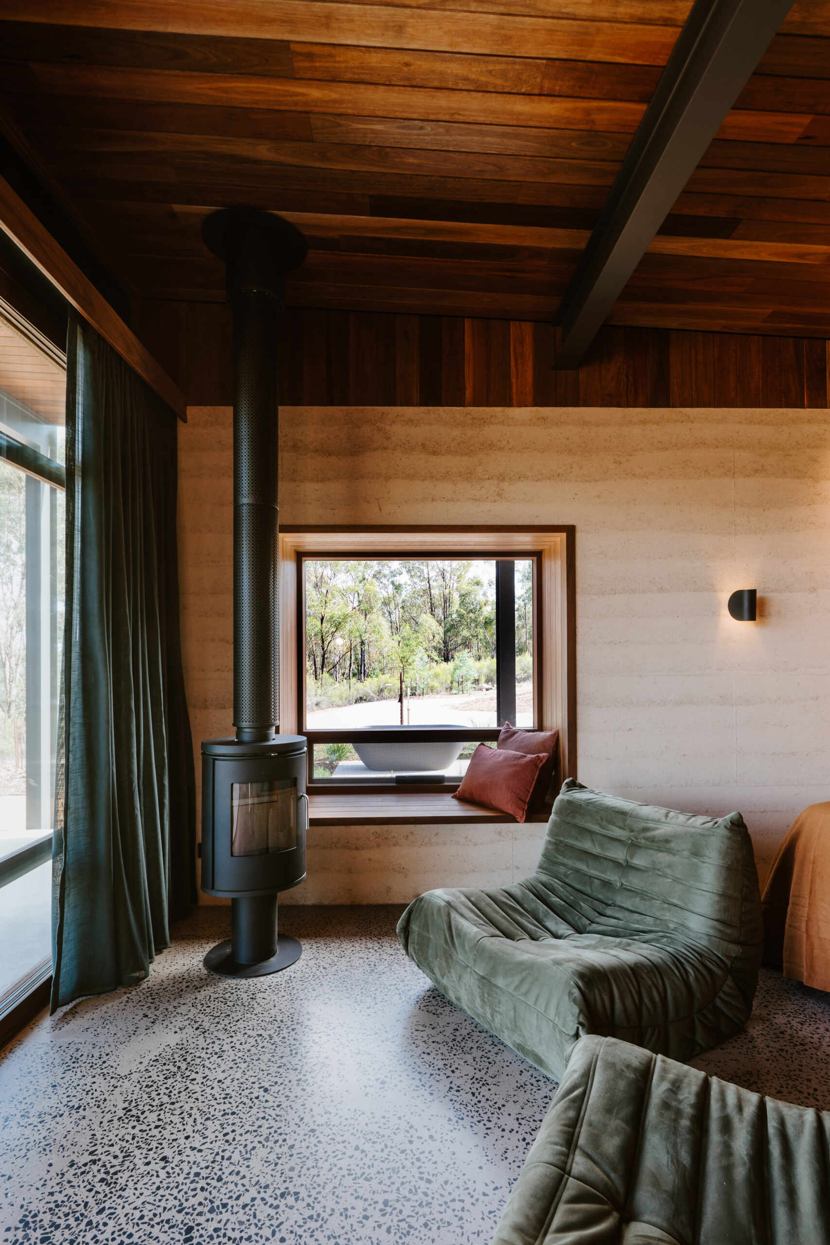 A contemporary off-grid cabin in Mudgee with an open-plan layout that combines the living area, bedroom, and kitchen into one light-filled space. Rammed earth walls, warm timber finishes, a minimalist bathroom, and an outdoor bathtub overlooking the landscape create a relaxing vineyard farm stay surrounded by Australian art and valley views.