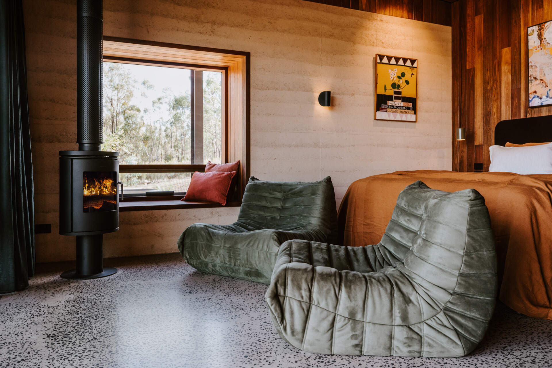 A contemporary off-grid cabin in Mudgee with an open-plan layout that combines the living area, bedroom, and kitchen into one light-filled space. Rammed earth walls, warm timber finishes, a minimalist bathroom, and an outdoor bathtub overlooking the landscape create a relaxing vineyard farm stay surrounded by Australian art and valley views.