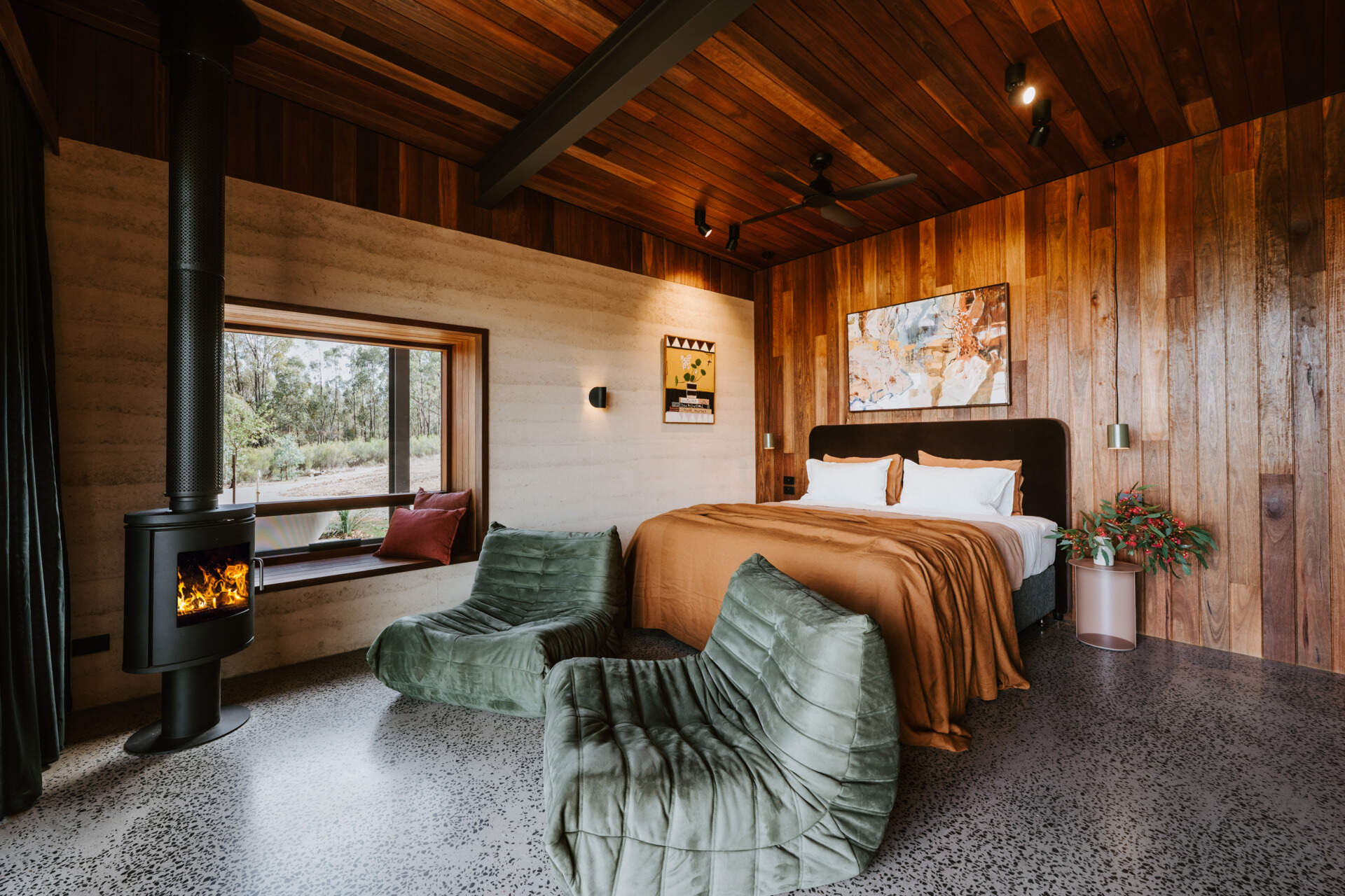 A contemporary off-grid cabin in Mudgee with an open-plan layout that combines the living area, bedroom, and kitchen into one light-filled space. Rammed earth walls, warm timber finishes, a minimalist bathroom, and an outdoor bathtub overlooking the landscape create a relaxing vineyard farm stay surrounded by Australian art and valley views.
