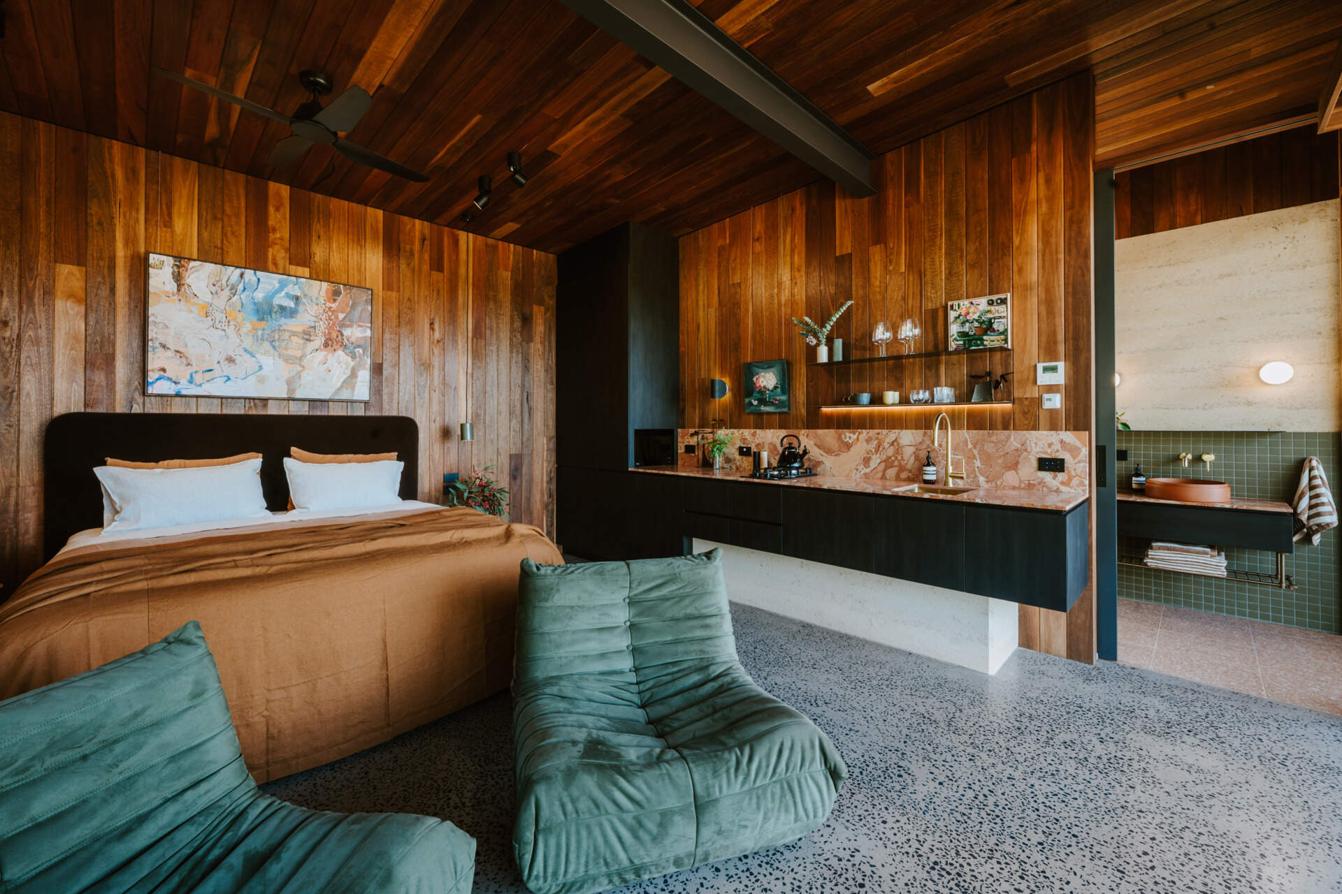 A contemporary off-grid cabin in Mudgee with an open-plan layout that combines the living area, bedroom, and kitchen into one light-filled space. Rammed earth walls, warm timber finishes, a minimalist bathroom, and an outdoor bathtub overlooking the landscape create a relaxing vineyard farm stay surrounded by Australian art and valley views.
