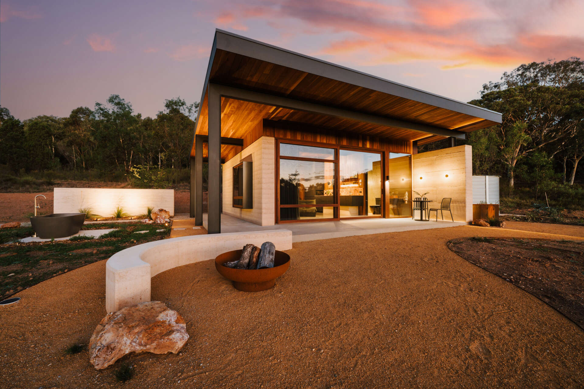 A contemporary off-grid cabin in Mudgee with an open-plan layout that combines the living area, bedroom, and kitchen into one light-filled space. Rammed earth walls, warm timber finishes, a minimalist bathroom, and an outdoor bathtub overlooking the landscape create a relaxing vineyard farm stay surrounded by Australian art and valley views.