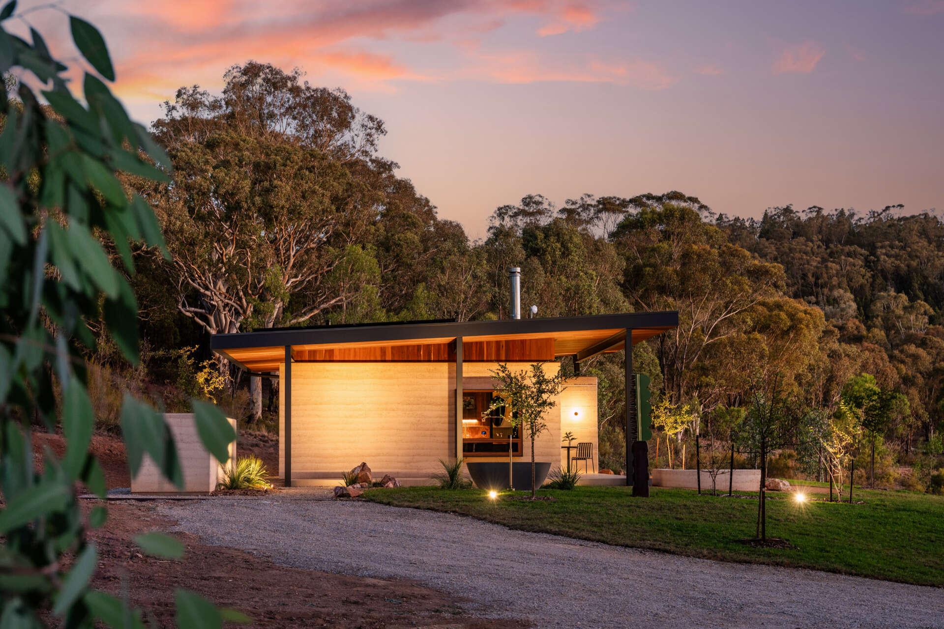 A contemporary off-grid cabin in Mudgee with an open-plan layout that combines the living area, bedroom, and kitchen into one light-filled space. Rammed earth walls, warm timber finishes, a minimalist bathroom, and an outdoor bathtub overlooking the landscape create a relaxing vineyard farm stay surrounded by Australian art and valley views.