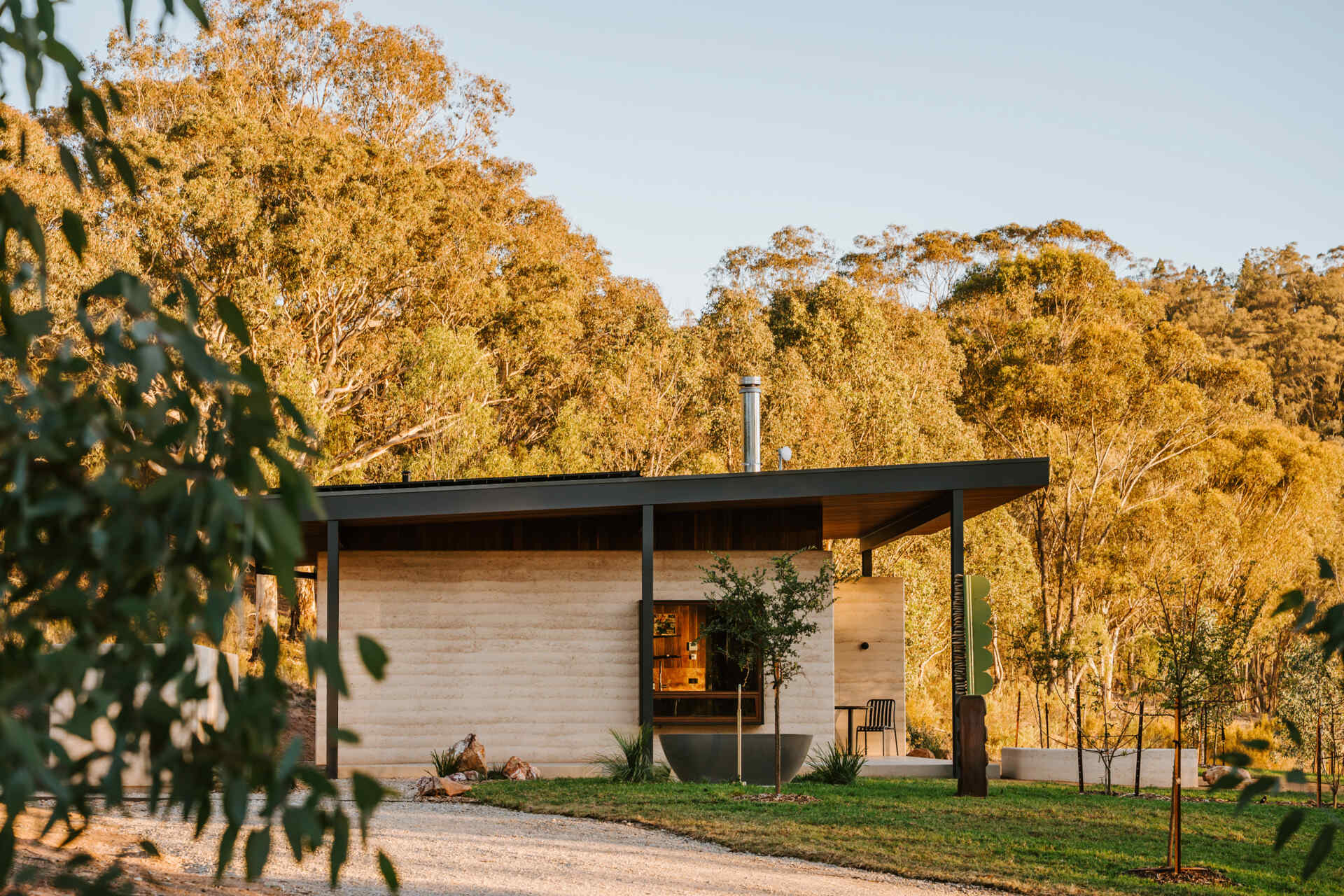 A contemporary off-grid cabin in Mudgee with an open-plan layout that combines the living area, bedroom, and kitchen into one light-filled space. Rammed earth walls, warm timber finishes, a minimalist bathroom, and an outdoor bathtub overlooking the landscape create a relaxing vineyard farm stay surrounded by Australian art and valley views.
