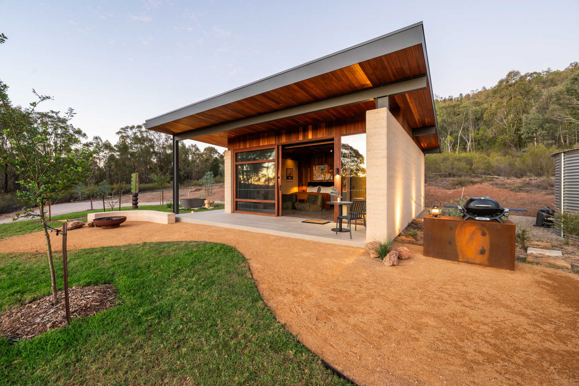 A contemporary off-grid cabin in Mudgee with an open-plan layout that combines the living area, bedroom, and kitchen into one light-filled space. Rammed earth walls, warm timber finishes, a minimalist bathroom, and an outdoor bathtub overlooking the landscape create a relaxing vineyard farm stay surrounded by Australian art and valley views.