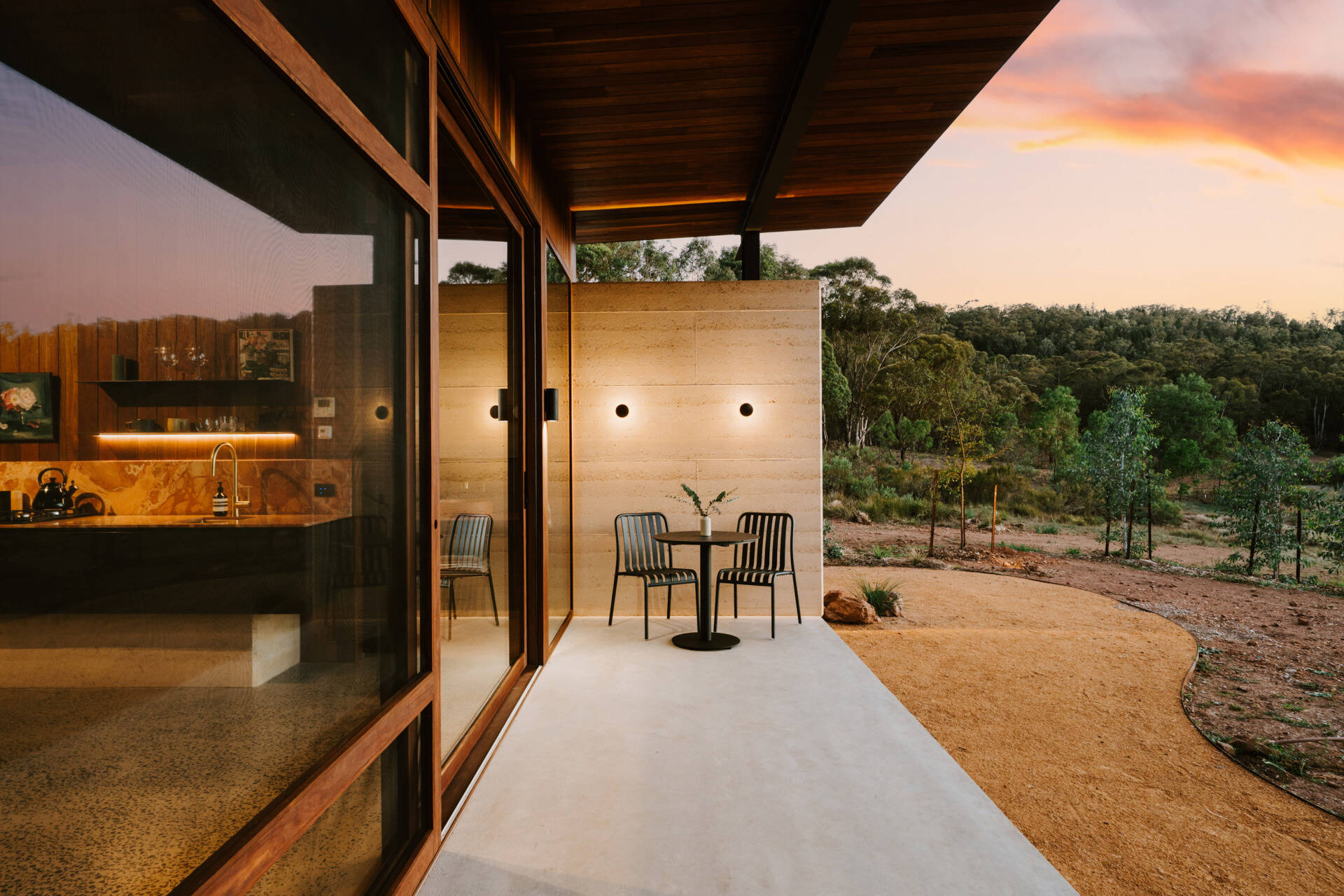 A contemporary off-grid cabin in Mudgee with an open-plan layout that combines the living area, bedroom, and kitchen into one light-filled space. Rammed earth walls, warm timber finishes, a minimalist bathroom, and an outdoor bathtub overlooking the landscape create a relaxing vineyard farm stay surrounded by Australian art and valley views.