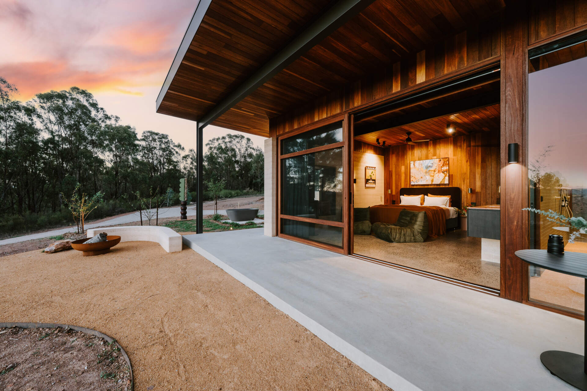 A contemporary off-grid cabin in Mudgee with an open-plan layout that combines the living area, bedroom, and kitchen into one light-filled space. Rammed earth walls, warm timber finishes, a minimalist bathroom, and an outdoor bathtub overlooking the landscape create a relaxing vineyard farm stay surrounded by Australian art and valley views.