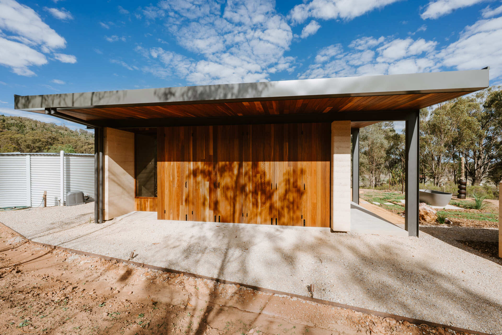A contemporary off-grid cabin in Mudgee with an open-plan layout that combines the living area, bedroom, and kitchen into one light-filled space. Rammed earth walls, warm timber finishes, a minimalist bathroom, and an outdoor bathtub overlooking the landscape create a relaxing vineyard farm stay surrounded by Australian art and valley views.