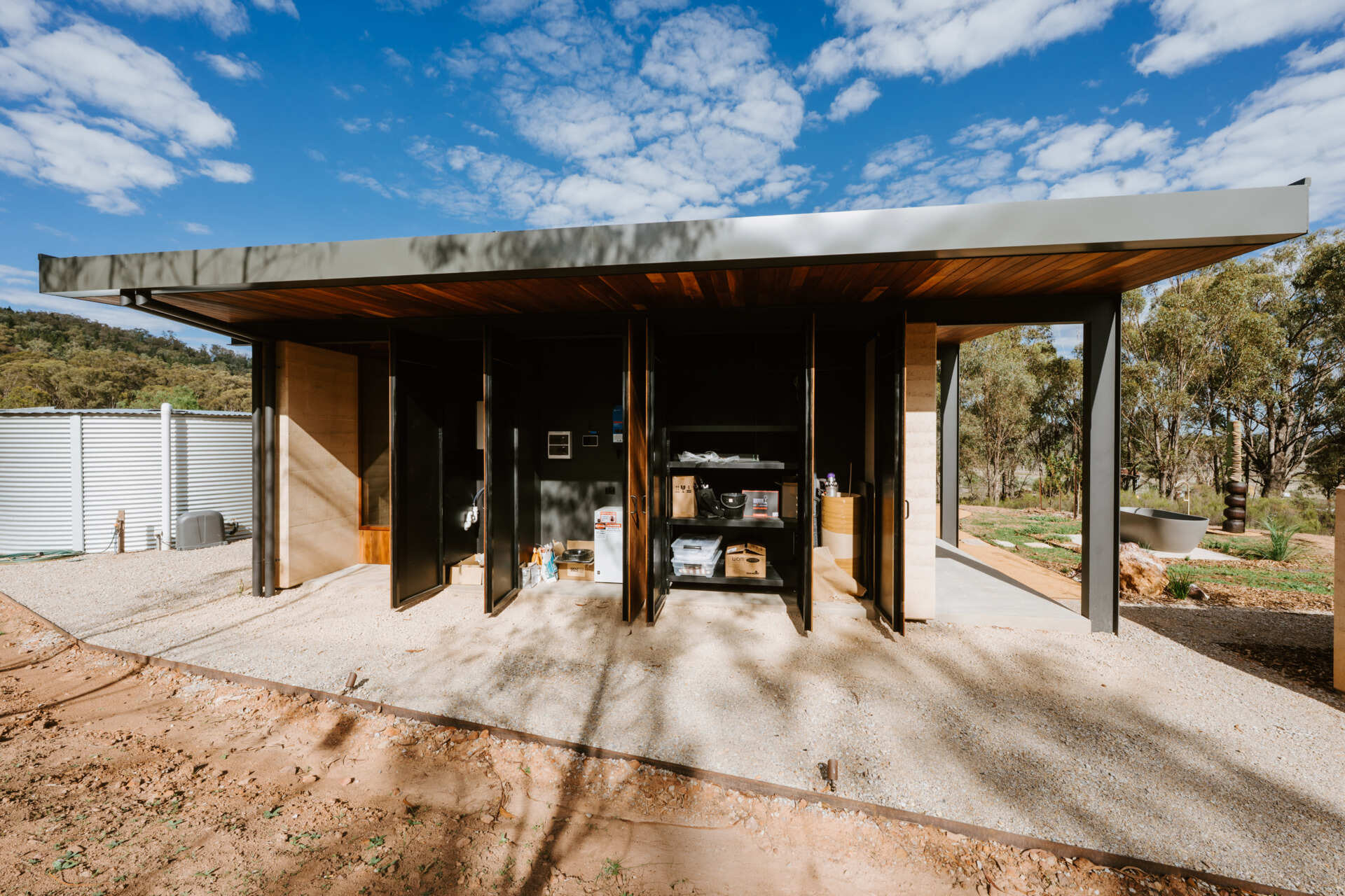 A contemporary off-grid cabin in Mudgee with an open-plan layout that combines the living area, bedroom, and kitchen into one light-filled space. Rammed earth walls, warm timber finishes, a minimalist bathroom, and an outdoor bathtub overlooking the landscape create a relaxing vineyard farm stay surrounded by Australian art and valley views.