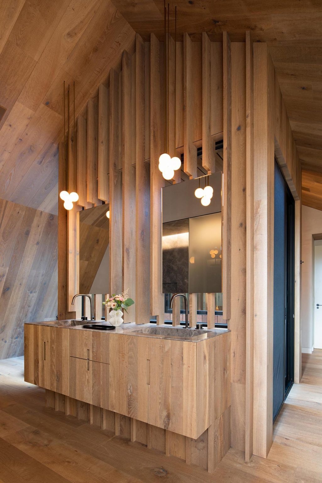 The en-suite bathroom continues the warm material palette with a wood vanity that blends seamlessly into the wall behind it. Vertical detailing emphasizes the height of the ceiling, while a freestanding bathtub and minimalist black faucet complete the serene space.