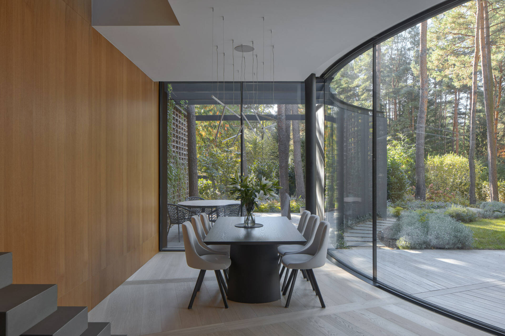 Inside House with a Glade, curved glass walls surround bright living and dining spaces with panoramic forest views. Natural light, oak flooring, and sweeping geometry define the home's interior atmosphere.