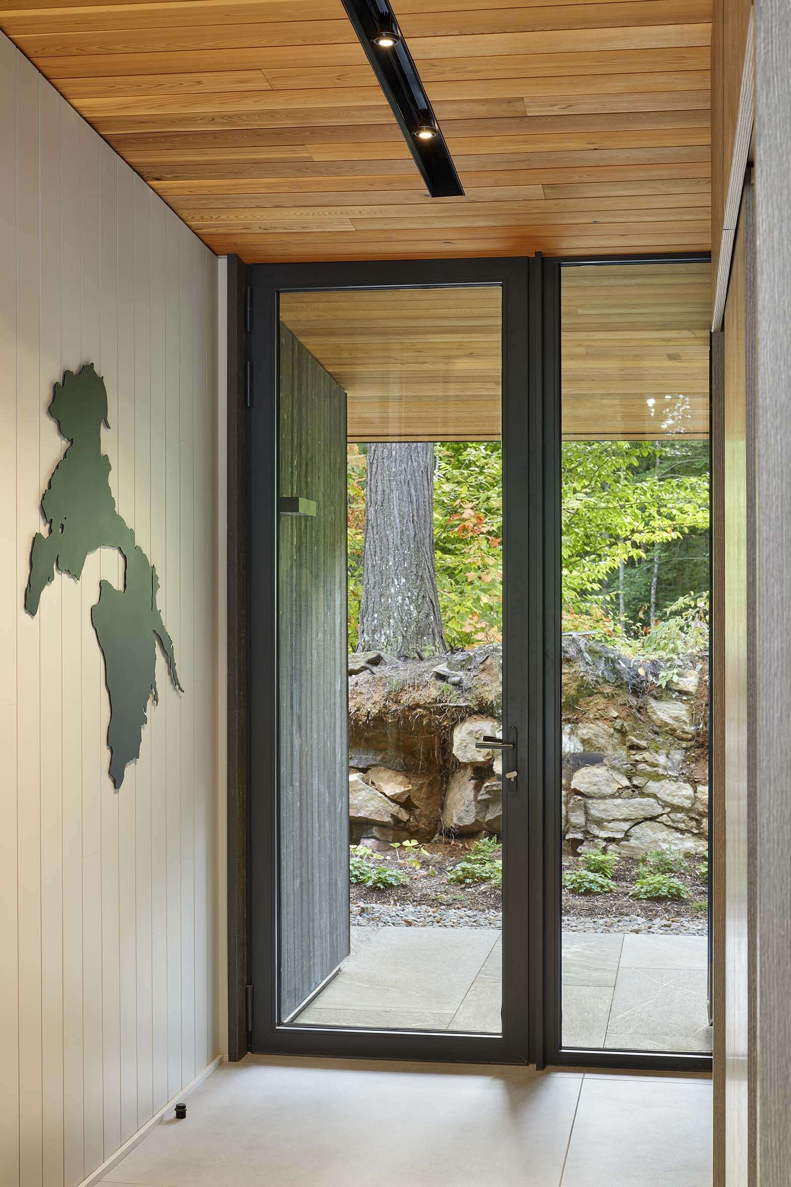 The entry is recessed and tucked behind the excavated embankment, creating a concealed arrival sequence rather than a prominent front approach. A blackened steel cut-out map of the lake is positioned at the entrance, referencing the site in a crafted detail.