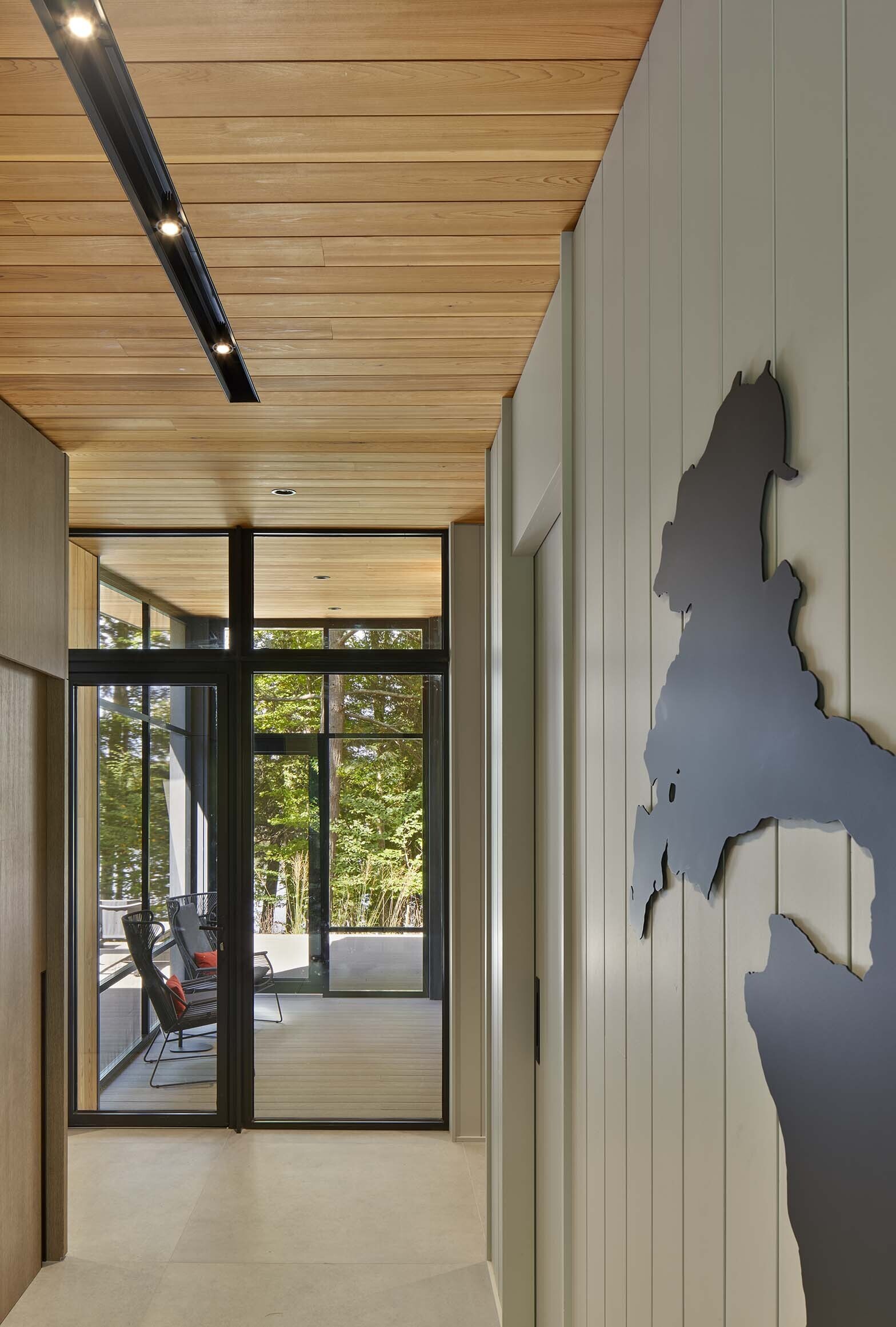 The entry is recessed and tucked behind the excavated embankment, creating a concealed arrival sequence rather than a prominent front approach. A blackened steel cut-out map of the lake is positioned at the entrance, referencing the site in a crafted detail.