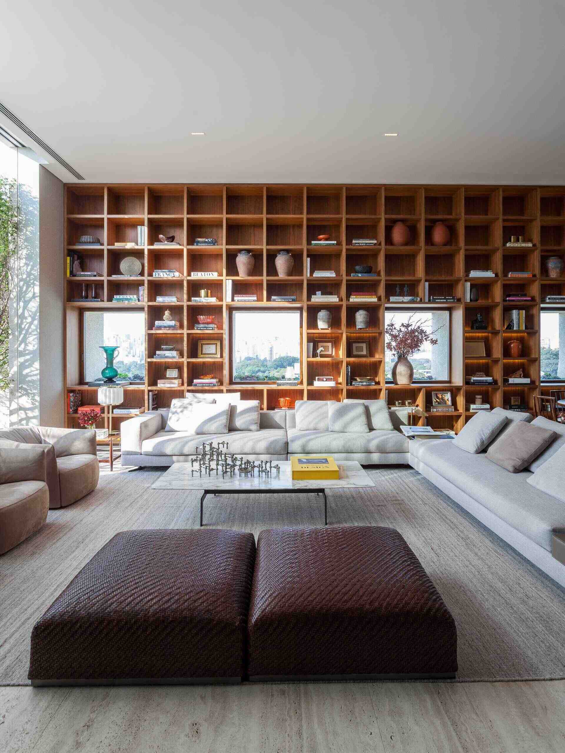 A stunning double-height living room in São Paulo features a floor-to-ceiling bookshelf wall that frames city views like artwork. Custom millwork, Brazilian design icons, and warm concealed lighting create a sophisticated and inviting interior.