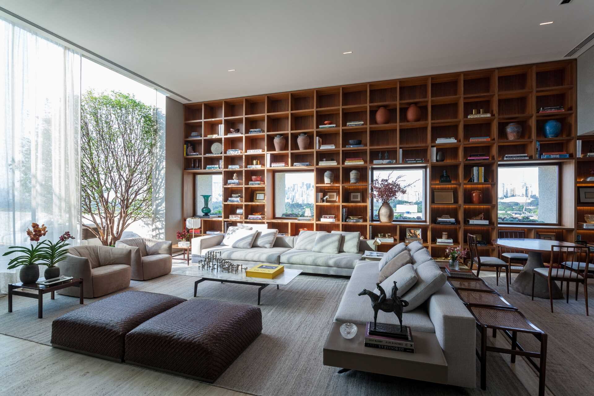 A stunning double-height living room in São Paulo features a floor-to-ceiling bookshelf wall that frames city views like artwork. Custom millwork, Brazilian design icons, and warm concealed lighting create a sophisticated and inviting interior.