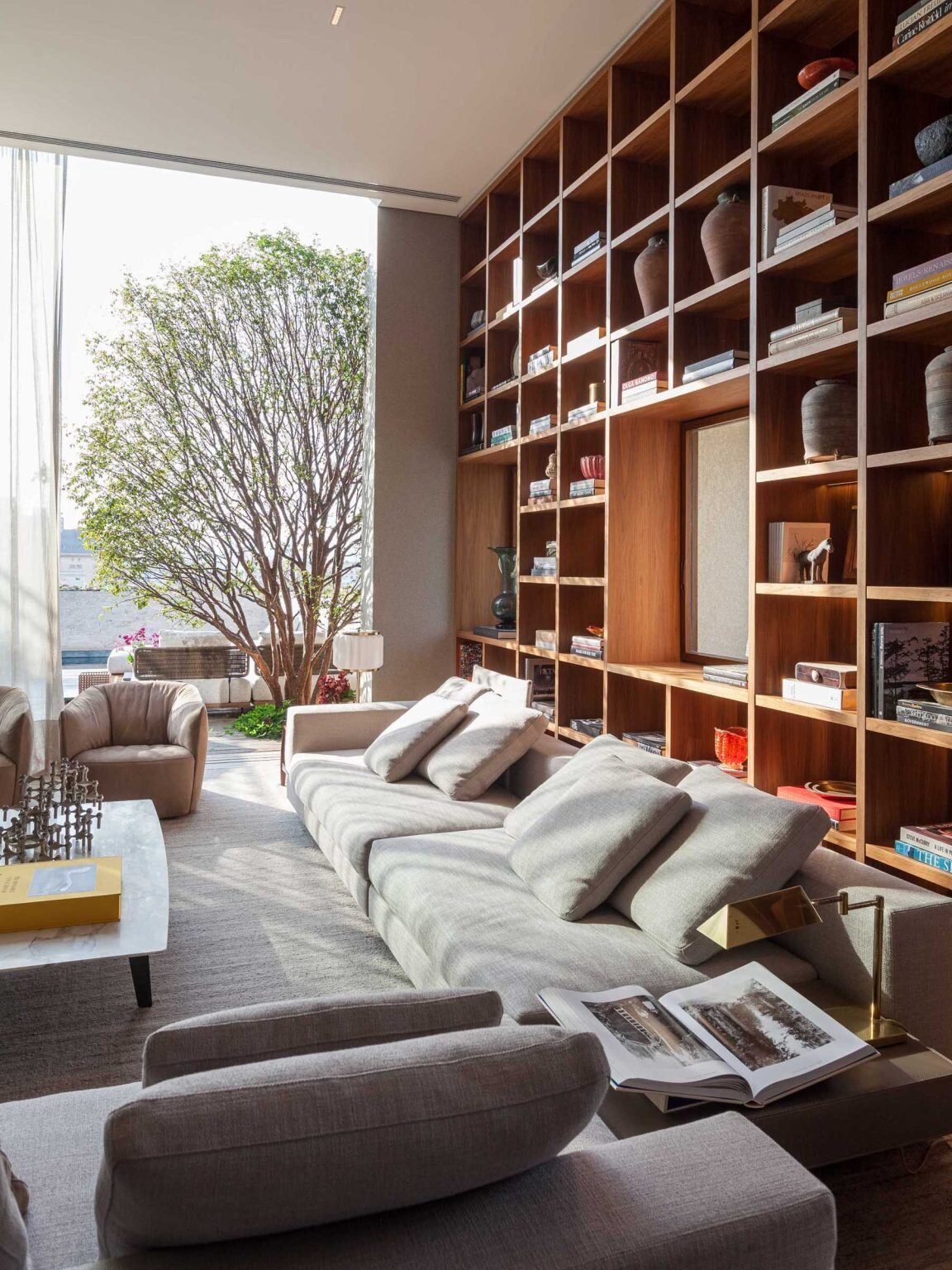 A stunning double-height living room in São Paulo features a floor-to-ceiling bookshelf wall that frames city views like artwork. Custom millwork, Brazilian design icons, and warm concealed lighting create a sophisticated and inviting interior.