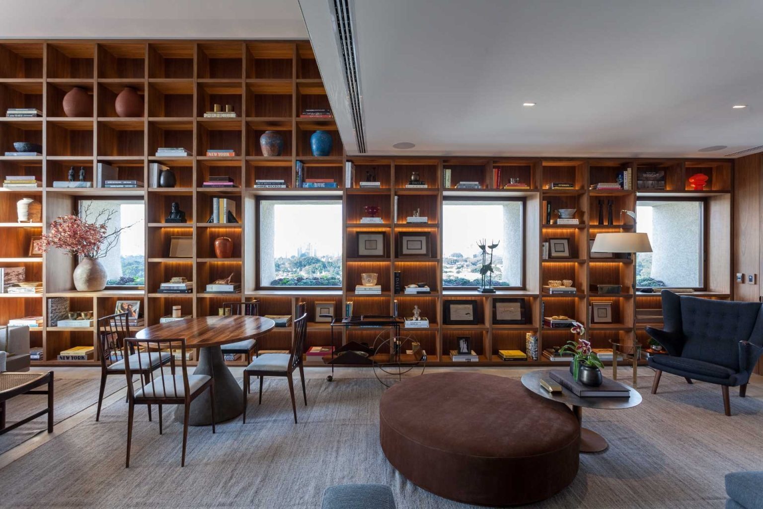 A stunning double-height living room in São Paulo features a floor-to-ceiling bookshelf wall that frames city views like artwork. Custom millwork, Brazilian design icons, and warm concealed lighting create a sophisticated and inviting interior.