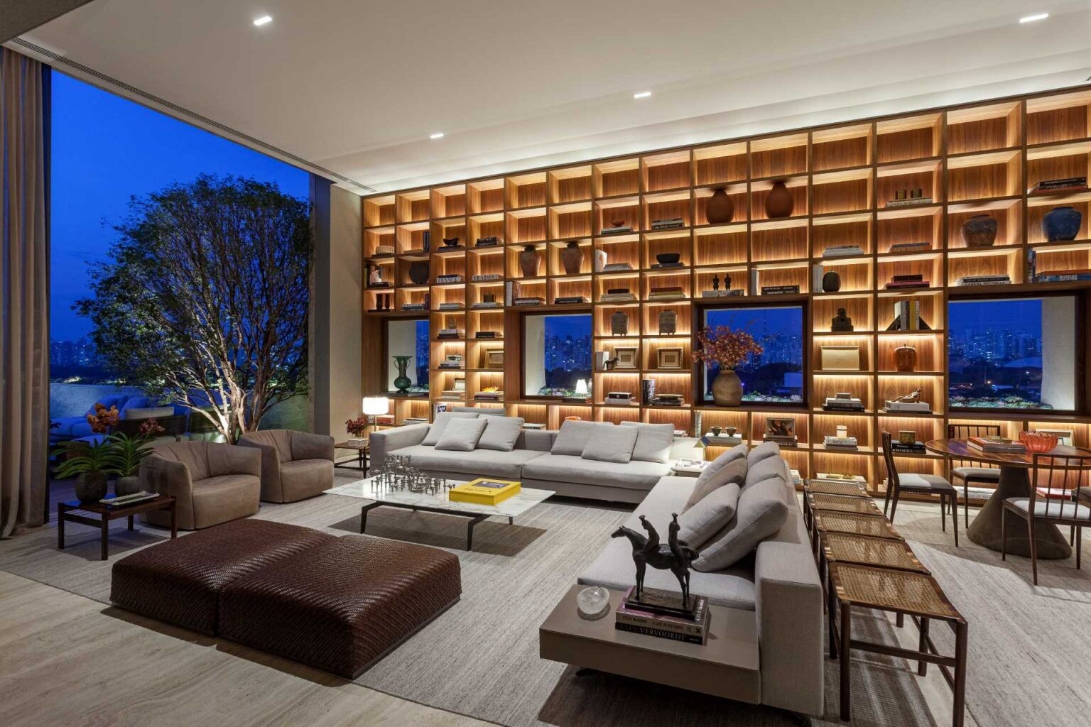 A stunning double-height living room in São Paulo features a floor-to-ceiling bookshelf wall that frames city views like artwork. Custom millwork, Brazilian design icons, and warm concealed lighting create a sophisticated and inviting interior.