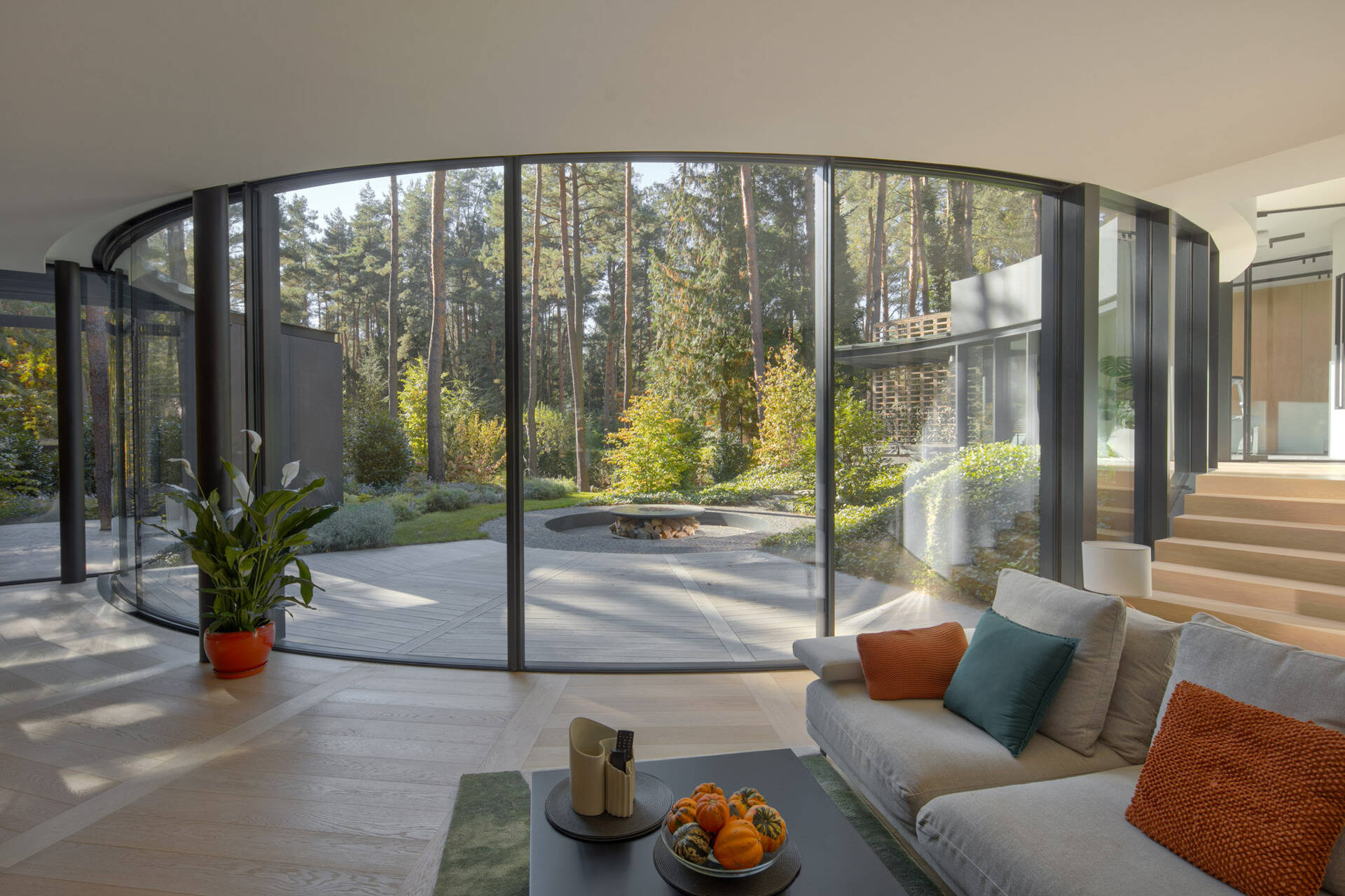 Inside House with a Glade, curved glass walls surround bright living and dining spaces with panoramic forest views. Natural light, oak flooring, and sweeping geometry define the home's interior atmosphere.