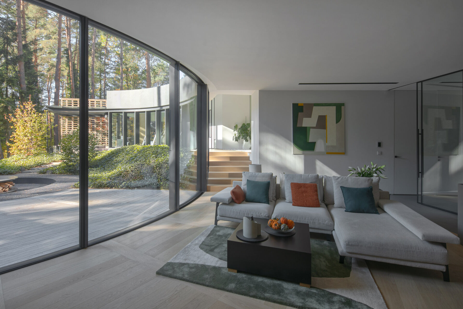 Inside House with a Glade, curved glass walls surround bright living and dining spaces with panoramic forest views. Natural light, oak flooring, and sweeping geometry define the home's interior atmosphere.