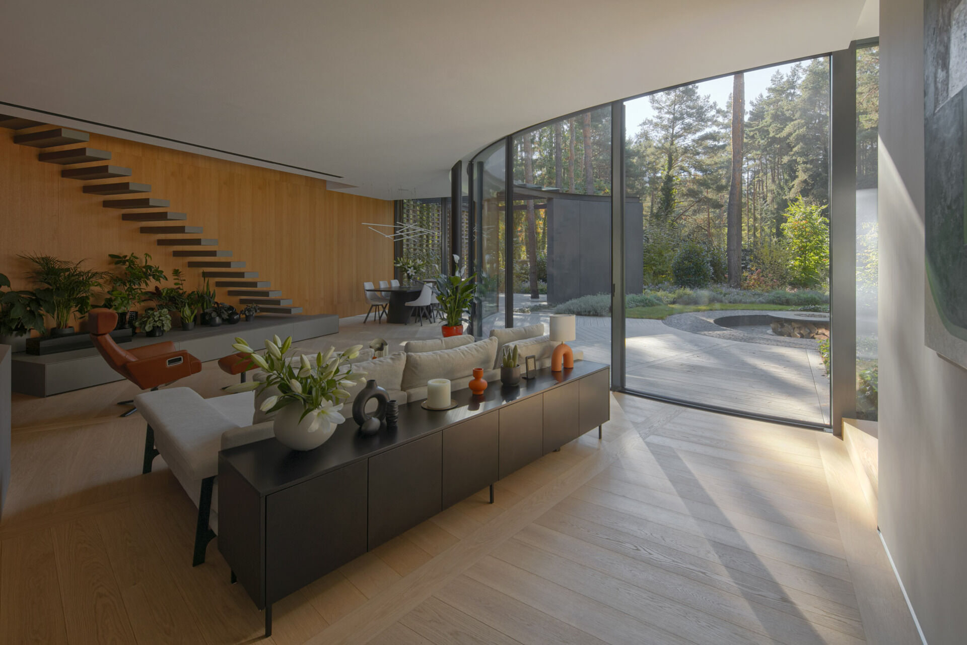 Inside House with a Glade, curved glass walls surround bright living and dining spaces with panoramic forest views. Natural light, oak flooring, and sweeping geometry define the home's interior atmosphere.
