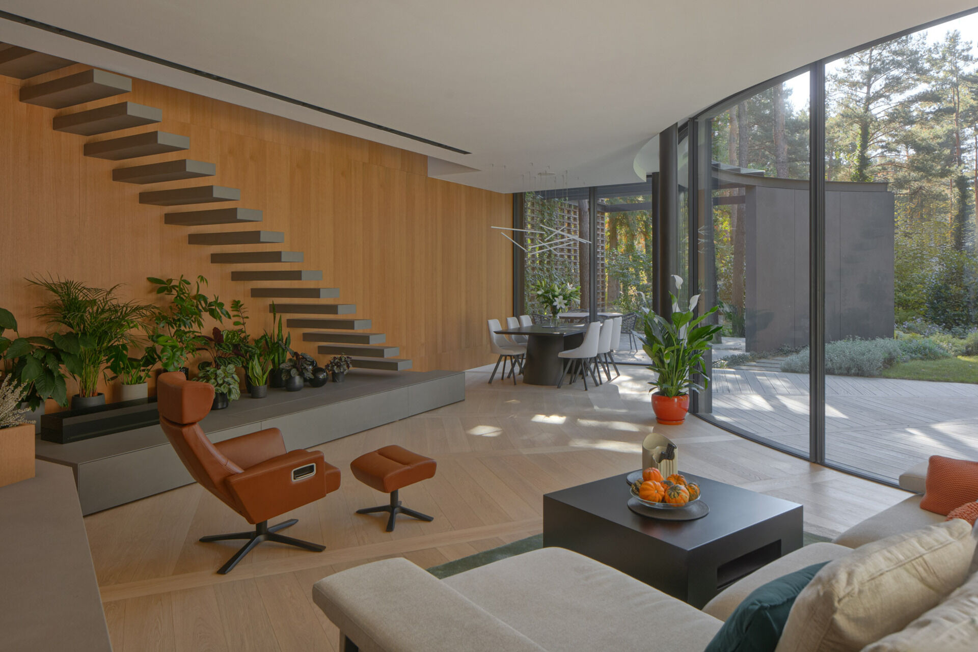 Inside House with a Glade, curved glass walls surround bright living and dining spaces with panoramic forest views. Natural light, oak flooring, and sweeping geometry define the home's interior atmosphere.