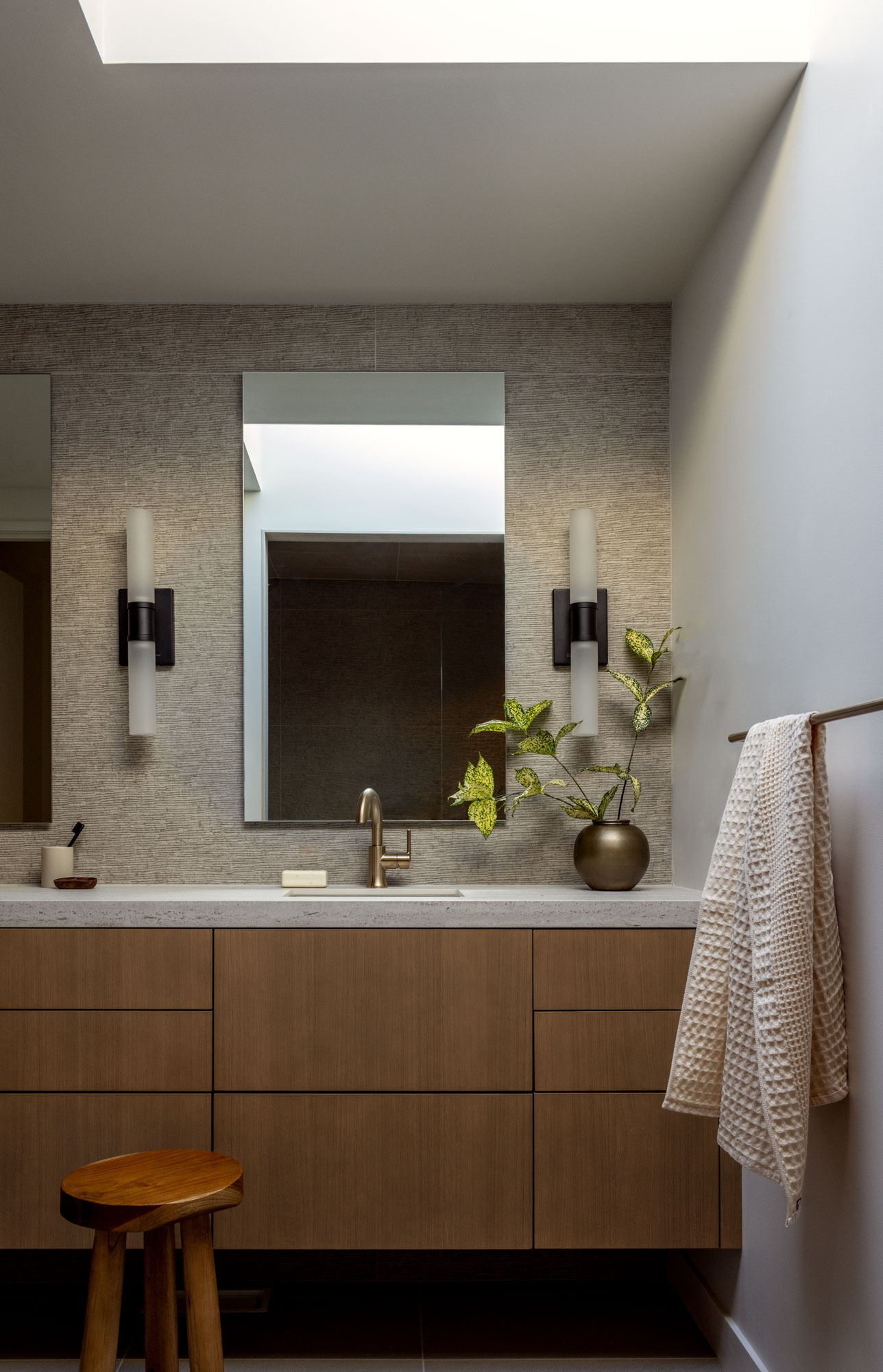 Natural light from the skylight brightens the bathroom, highlighting the textured wall tile. A warm wood vanity, brushed brass faucet, and simple stone countertop bring natural materials together in a clean, timeless palette that complements the home's understated aesthetic.
