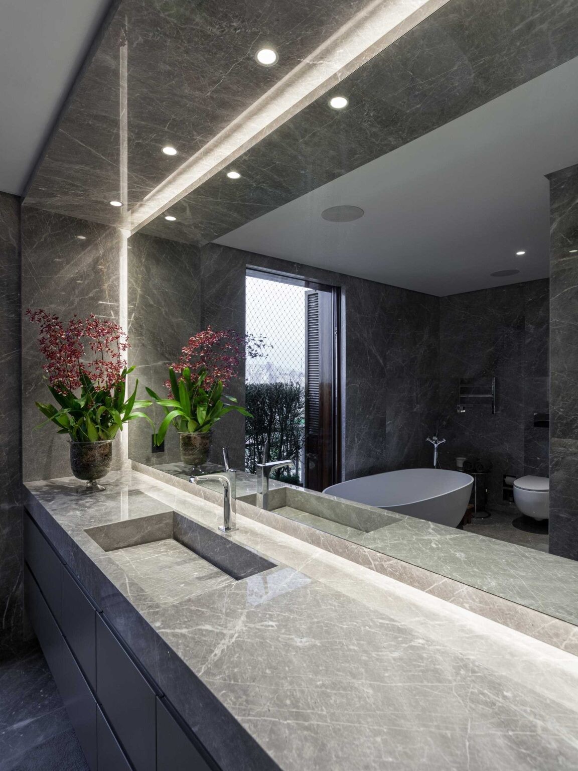 This modern bathroom introduces a built-in stone sink, walk-in shower with integrated bench and niche, and a freestanding bathtub placed beside the window. Here, dramatic Verde Guatemala marble wraps the walls and vanity, creating a bold visual statement.