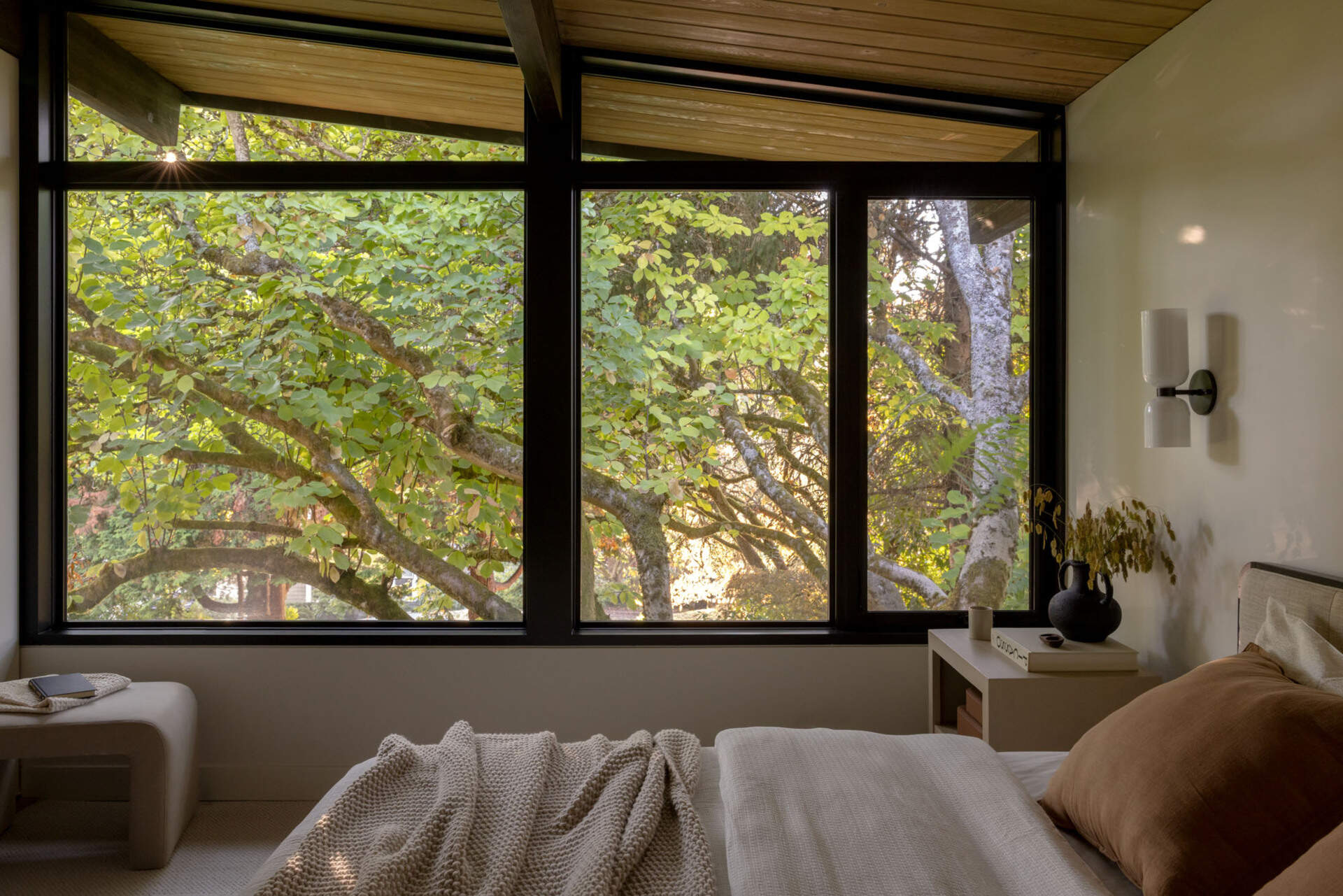 Large windows frame leafy views in this peaceful bedroom, where exposed steel beams, timber ceilings, and soft neutral finishes create a bright, calming space inspired by the surrounding landscape.