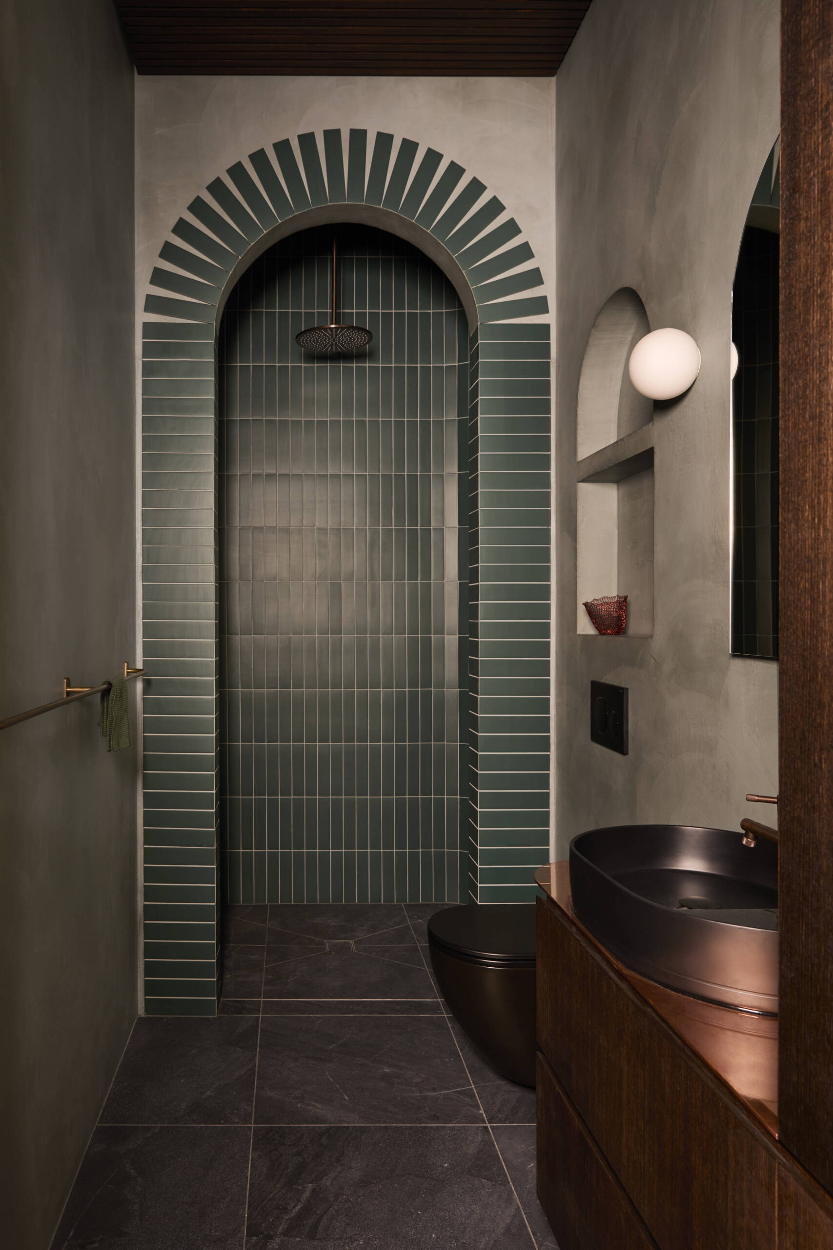 A moody bathroom with deep green tiles, an arched shower, warm timber cabinetry, and dark stone floors. This compact bathroom is packed with bold color, natural textures, and modern design inspiration.