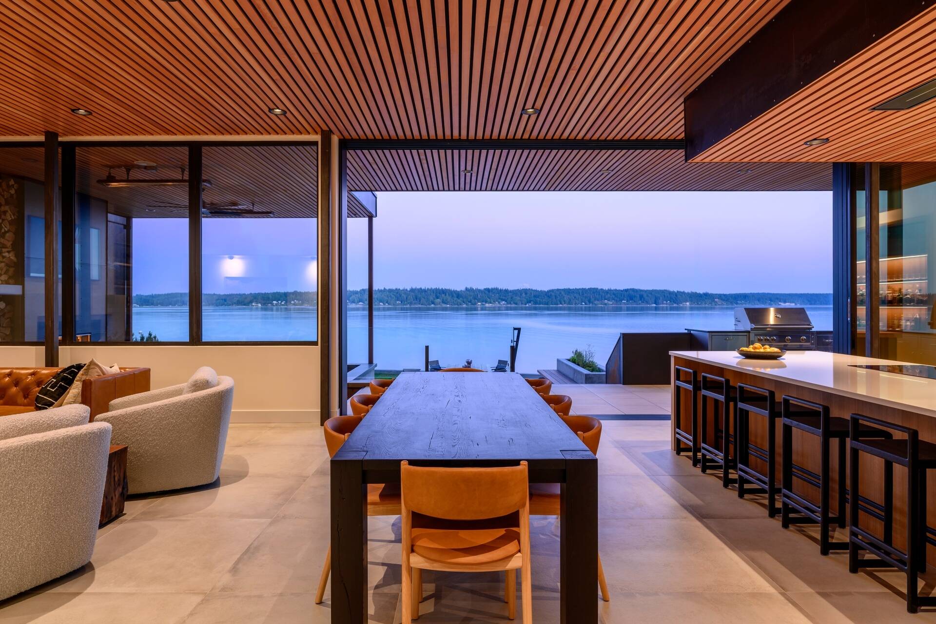 Discover a waterfront dining space where a long timber table, seamless indoor-outdoor living, and panoramic Puget Sound views create the perfect setting for entertaining.