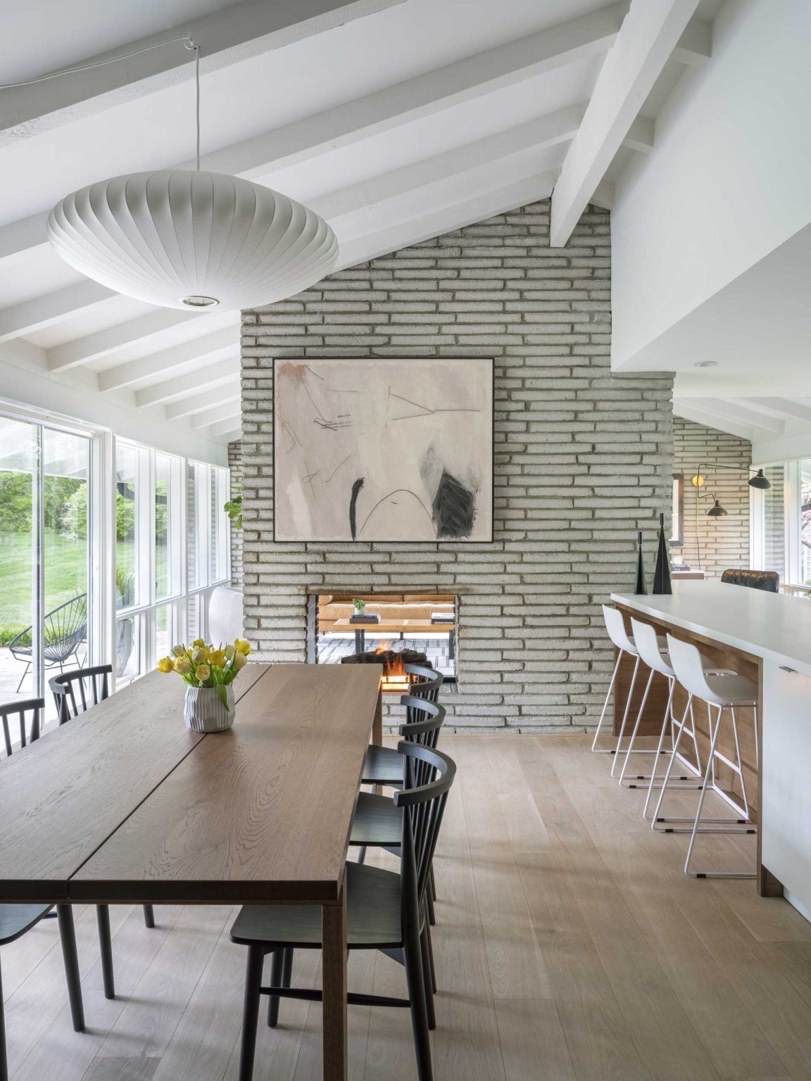 An original living room becomes a bright dining space centered around a preserved double-sided brick fireplace. Open views and mid-century furnishings help connect the home's updated interiors.