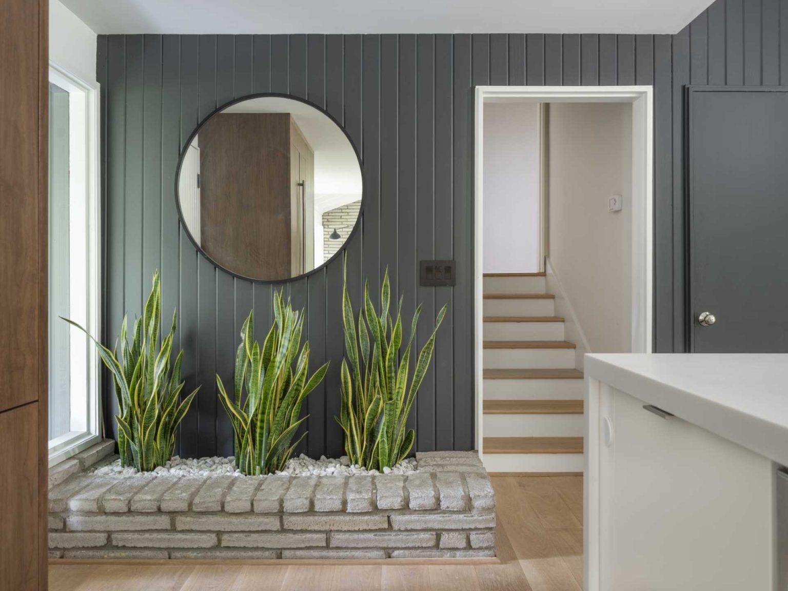 See how a dark mid-century entryway was transformed with light wood floors, grey finishes, and a large round mirror, while an original brick planter remains as a nod to the home's history.