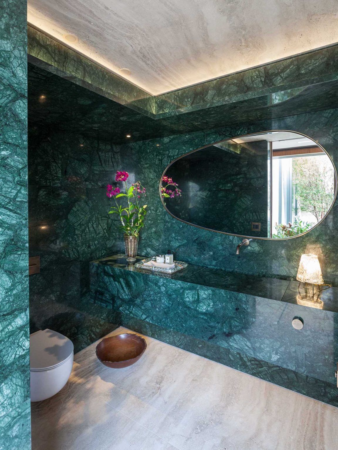 Measuring only four square meters, this powder room combines Verde Guatemala marble, Roman Travertine flooring, a carved stone sink, indirect lighting, and a brass-framed mirror to create a memorable experience.