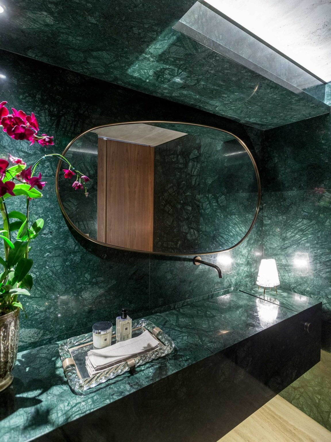 Measuring only four square meters, this powder room combines Verde Guatemala marble, Roman Travertine flooring, a carved stone sink, indirect lighting, and a brass-framed mirror to create a memorable experience.