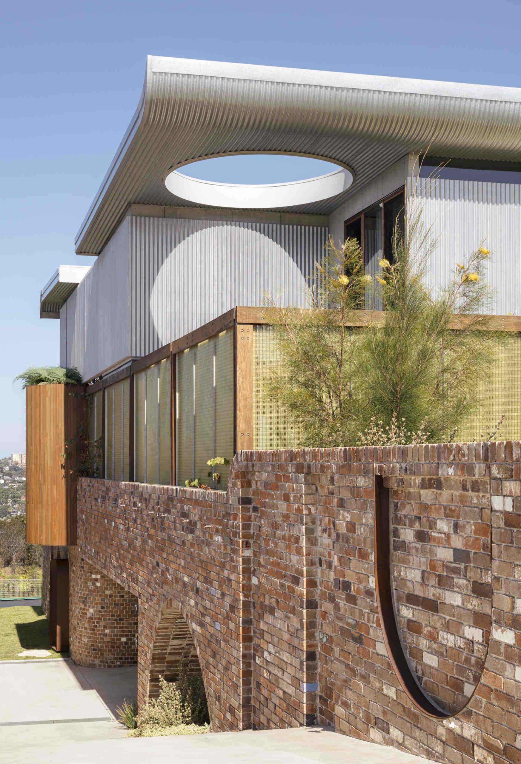 A stunning Sydney home featuring a dramatic butterfly roof, circular windows, curved brickwork, and translucent jade-green screens that create privacy without blocking light.